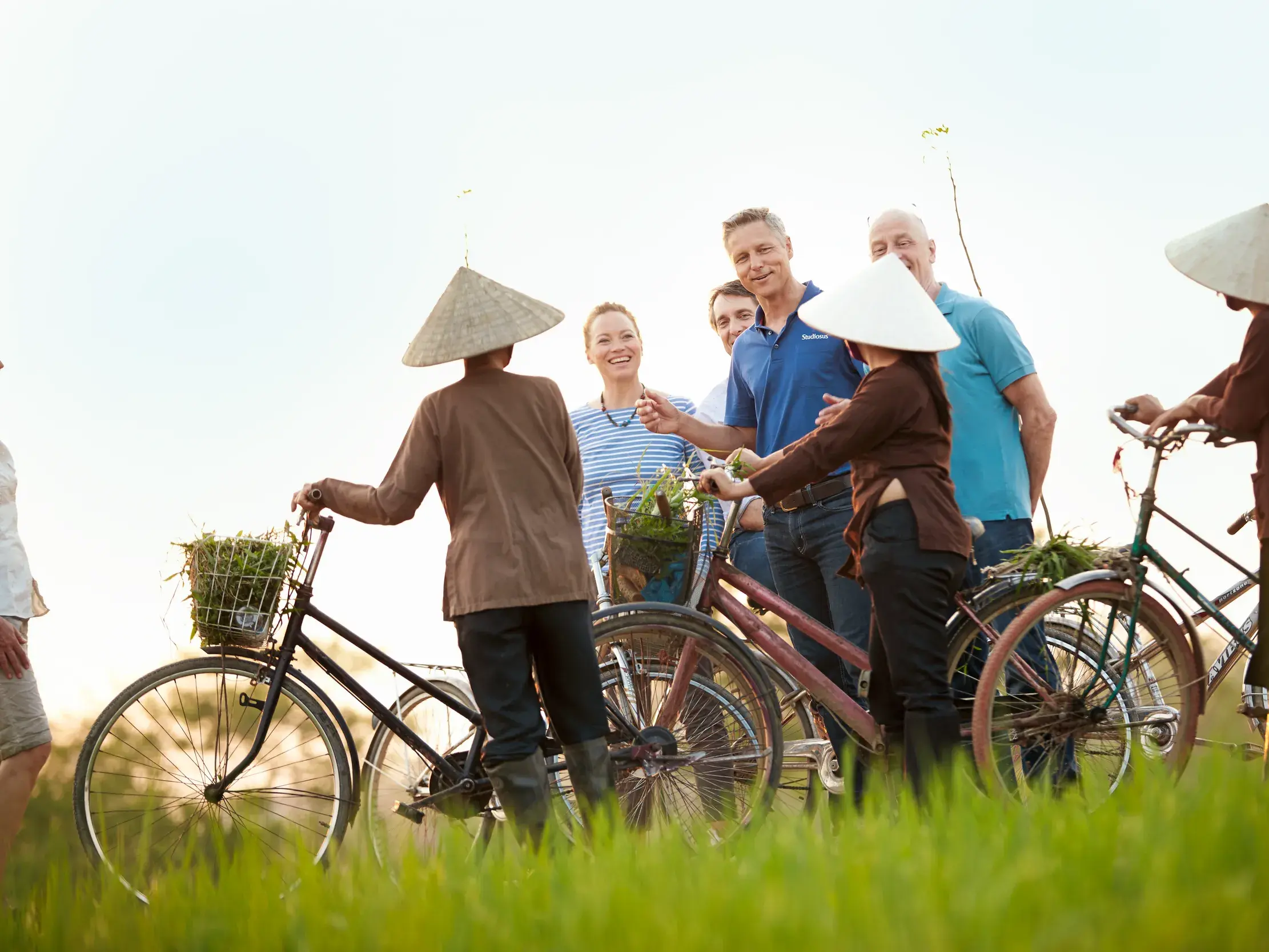 Eine Kleingruppe von Reisenden spricht mit Bäuerinnen in Kegelhüten während einer Radtour durch die grünen Felder von Tra Que.