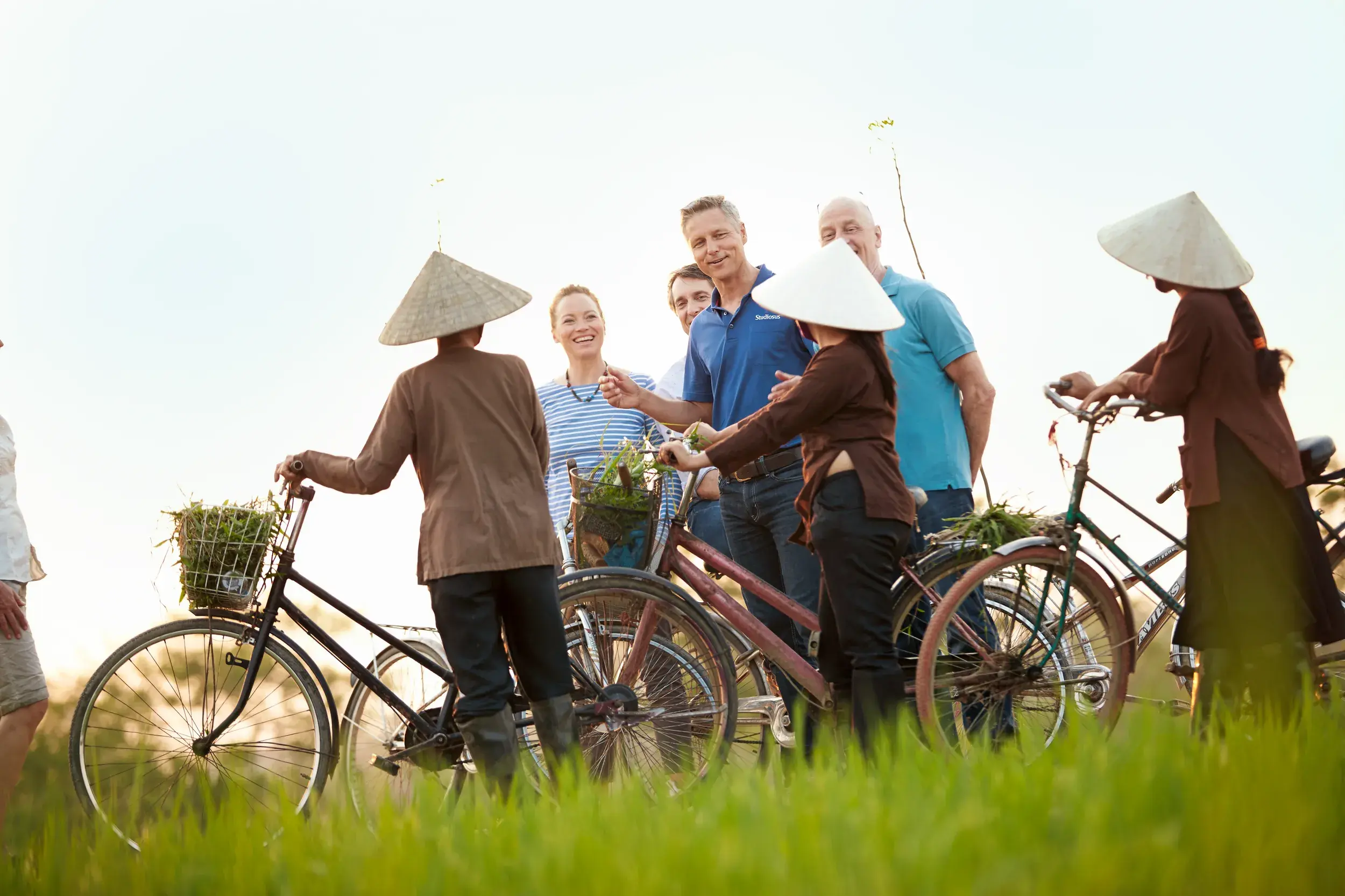 Eine Kleingruppe von Reisenden spricht mit Bäuerinnen in Kegelhüten während einer Radtour durch die grünen Felder von Tra Que.