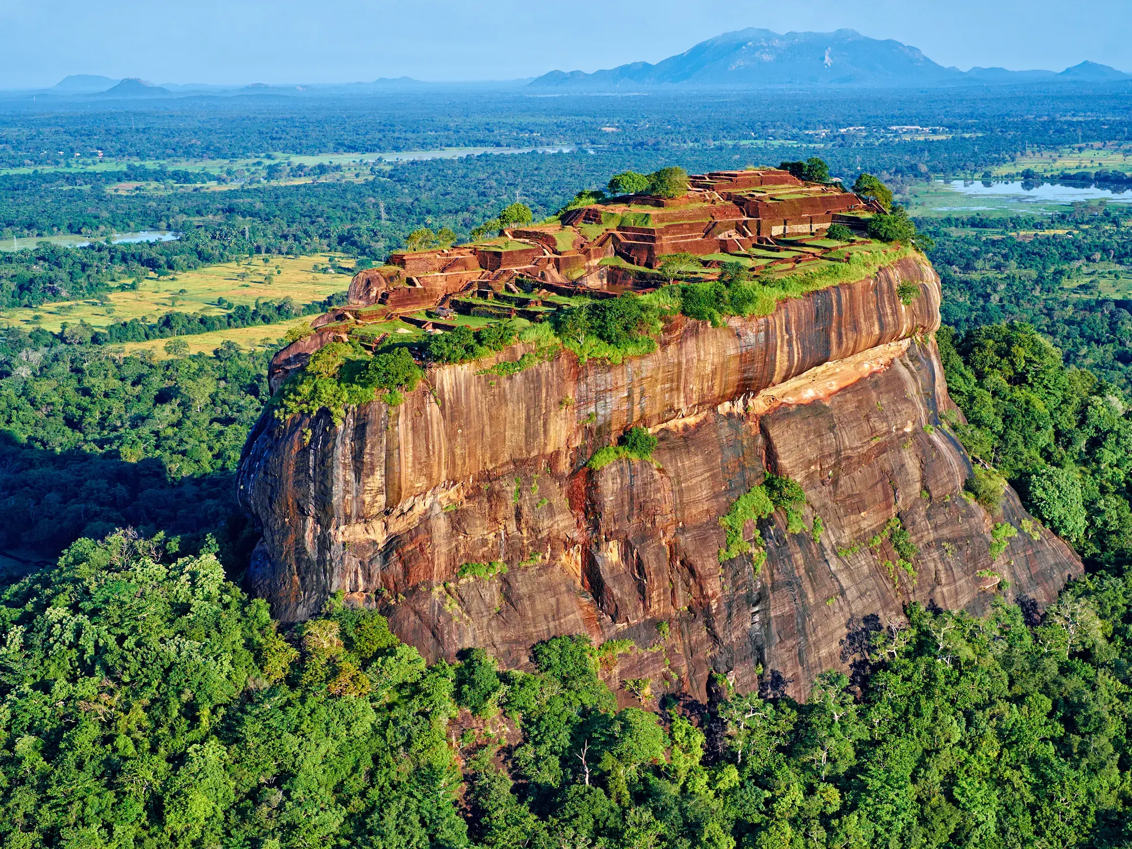 Luftaufnahme der historischen Felsenfestung Sigiriya, die aus der weiten, dschungelgrünen Ebene Sri Lankas emporragt.