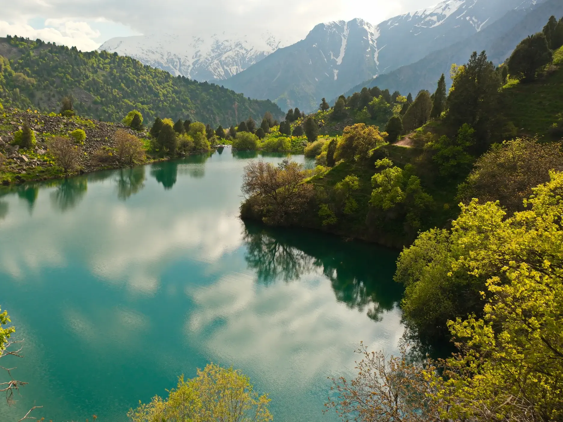 Ruhiger, smaragdgrüner Yashilkul-See in Tadschikistan, umgeben von grünen Hängen vor der Kulisse schneebedeckter Pamir-Gipfel.