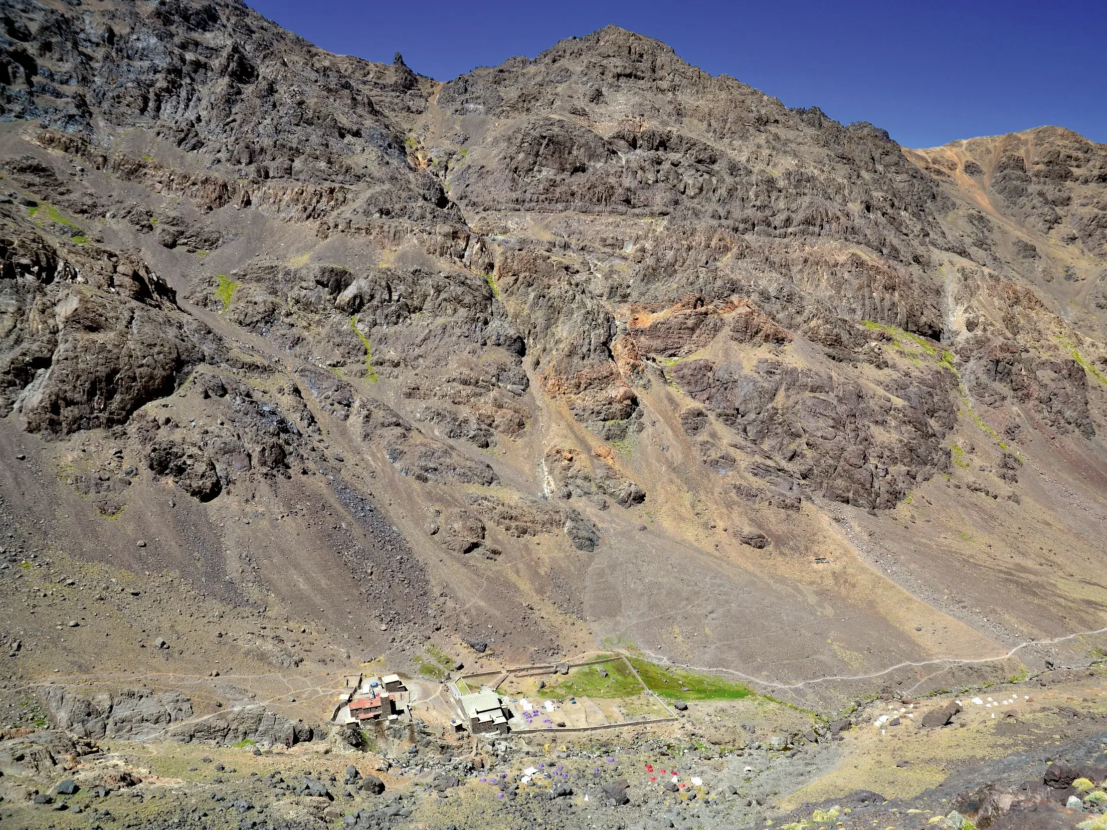 Das Toubkal Base Camp in einem weiten, kargen Tal im Hohen Atlas in Marokko. Ein schmaler Wanderpfad führt den Felshang hinauf.