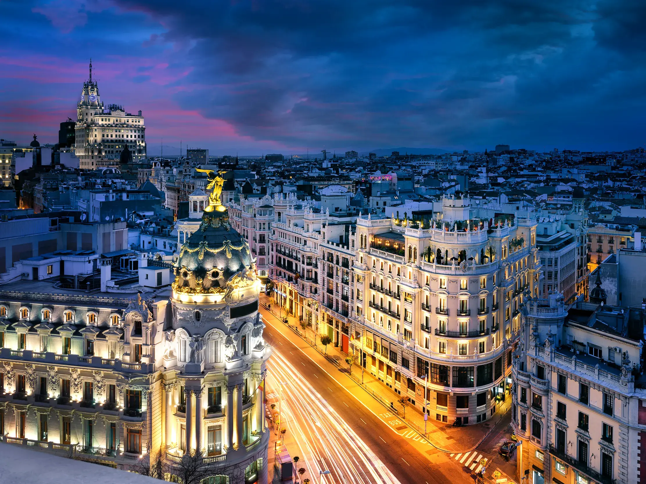 Blick auf die beleuchtete Gran Vía in Madrid in der Dämmerung mit dem Metrópolis-Gebäude und Lichtspuren des Verkehrs.