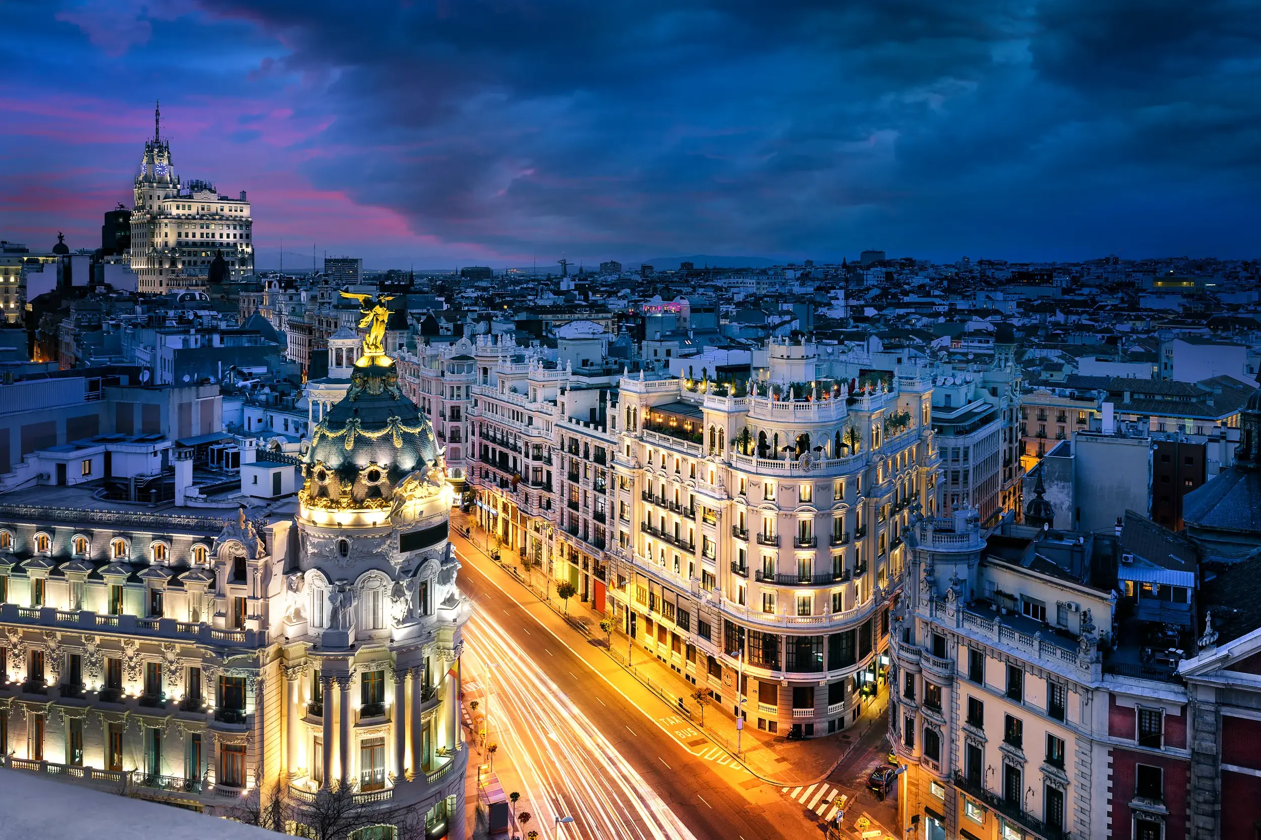 Blick auf die beleuchtete Gran Vía in Madrid in der Dämmerung mit dem Metrópolis-Gebäude und Lichtspuren des Verkehrs.