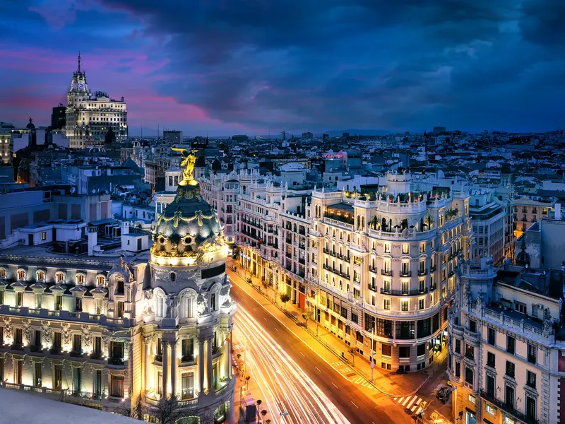 Blick auf die beleuchtete Gran Vía in Madrid in der Dämmerung mit dem Metrópolis-Gebäude und Lichtspuren des Verkehrs.