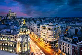 Blick auf die beleuchtete Gran Vía in Madrid in der Dämmerung mit dem Metrópolis-Gebäude und Lichtspuren des Verkehrs.