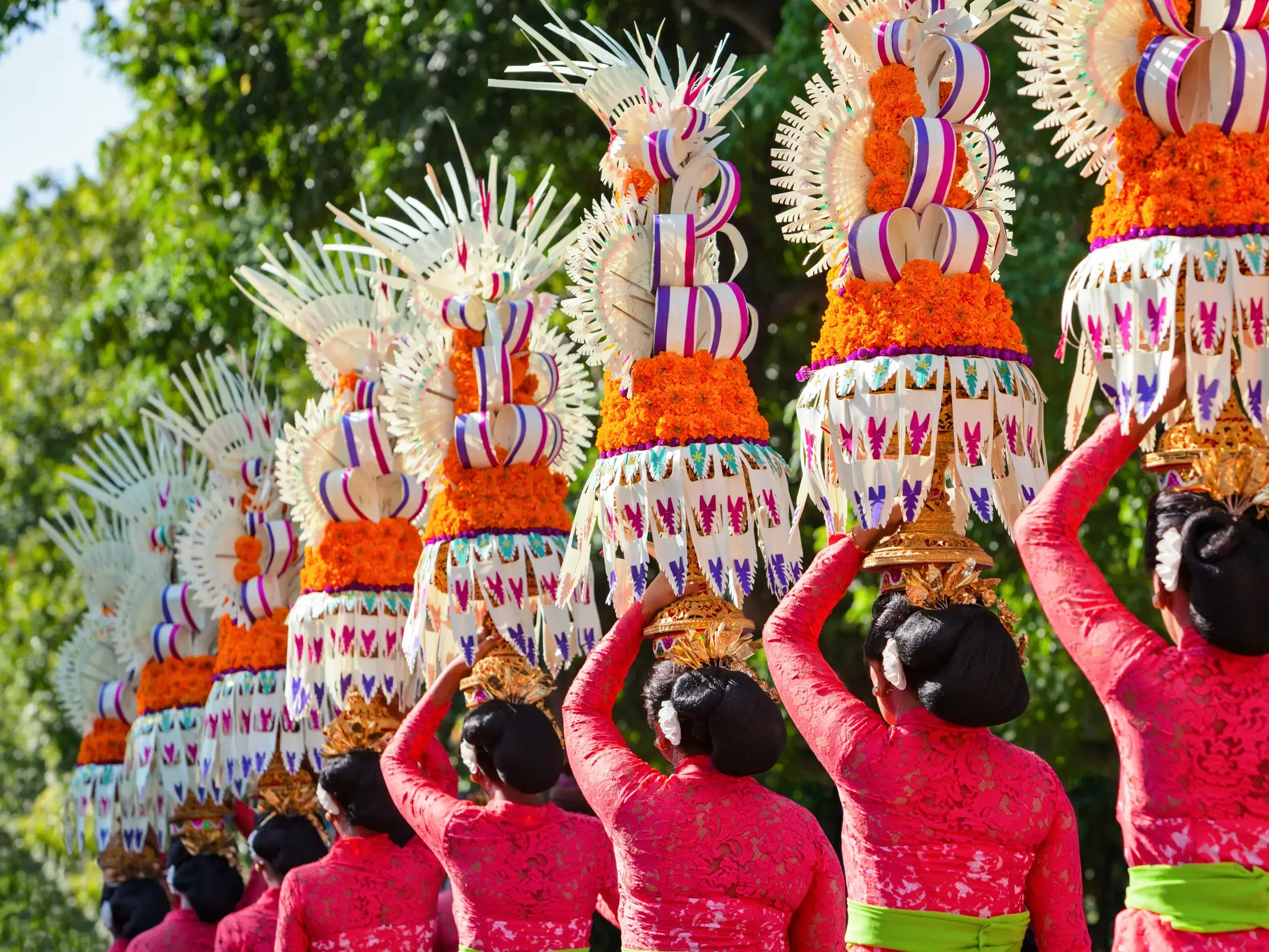 Rückansicht einer Gruppe balinesischer Frauen in roter Tracht, die bei einem Tempelfest auf Bali hohe, kunstvolle Opfergaben auf dem Kopf tragen.