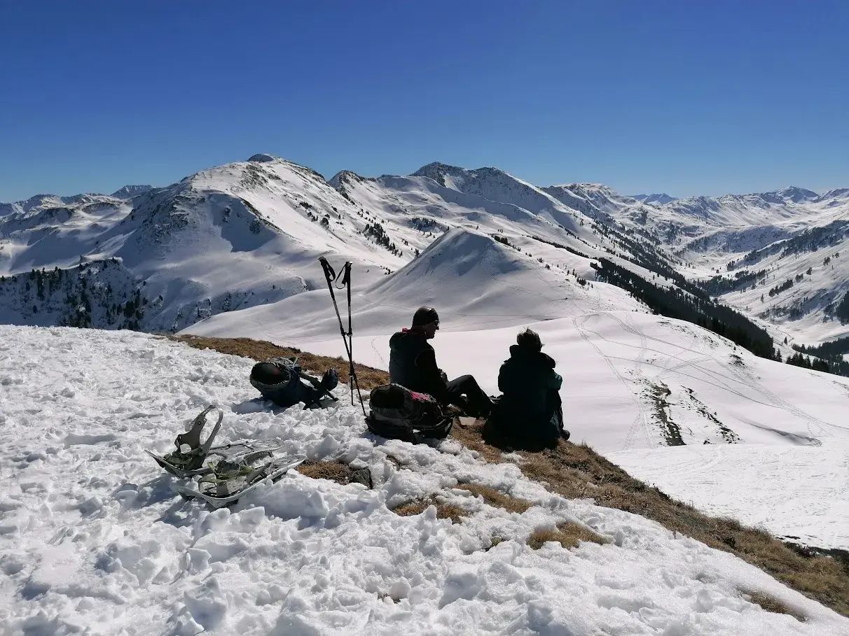 Zwei Wanderer genießen bei einer Gipfelrast mit Schneeschuhen den sonnigen Panoramablick auf die verschneiten Kitzbüheler Alpen.