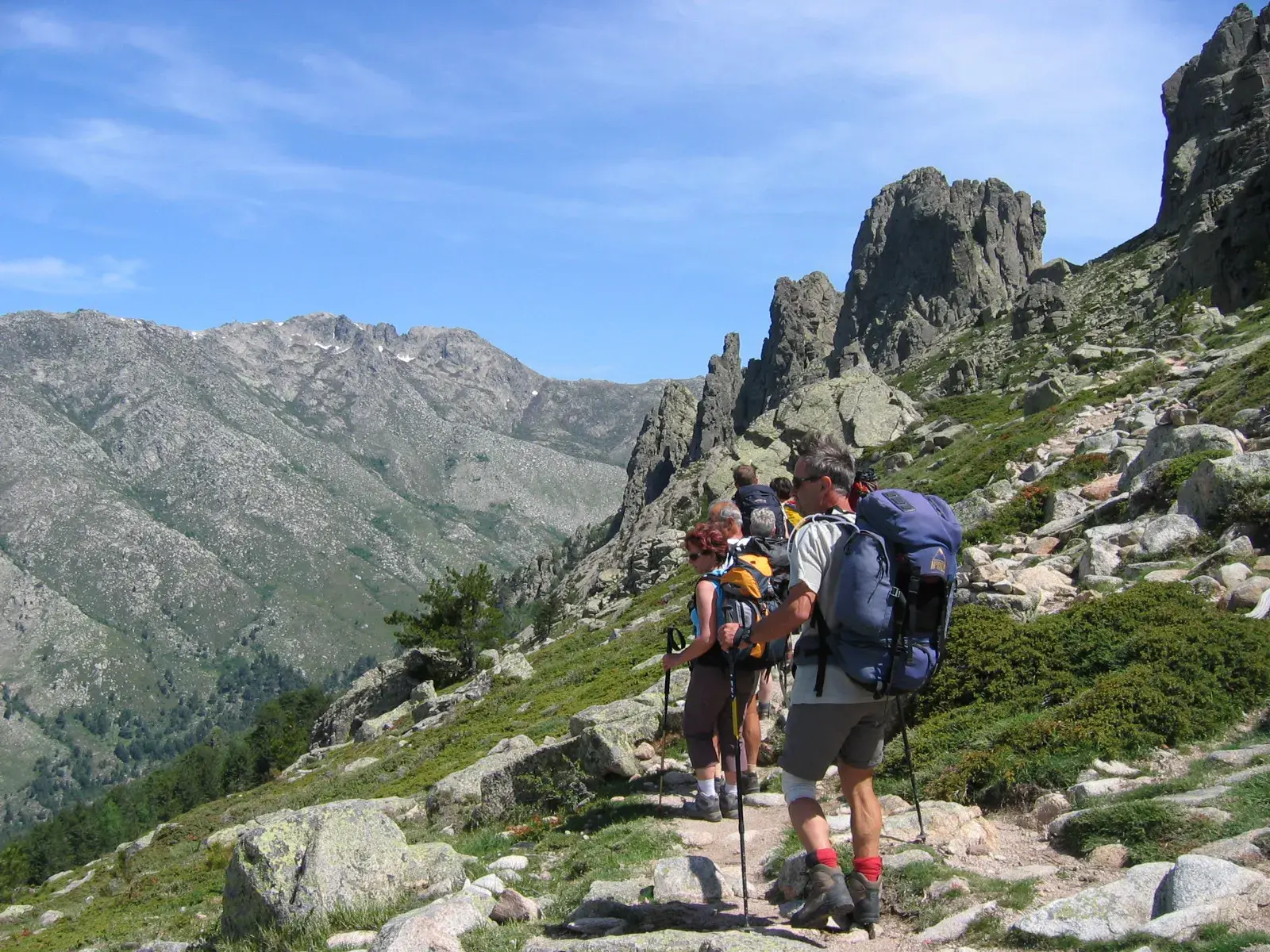 Eine kleine Wandergruppe mit Trekkingrucksäcken auf dem steinigen GR20-Pfad mit Blick auf die Felsnadeln des Bavella-Massivs.
