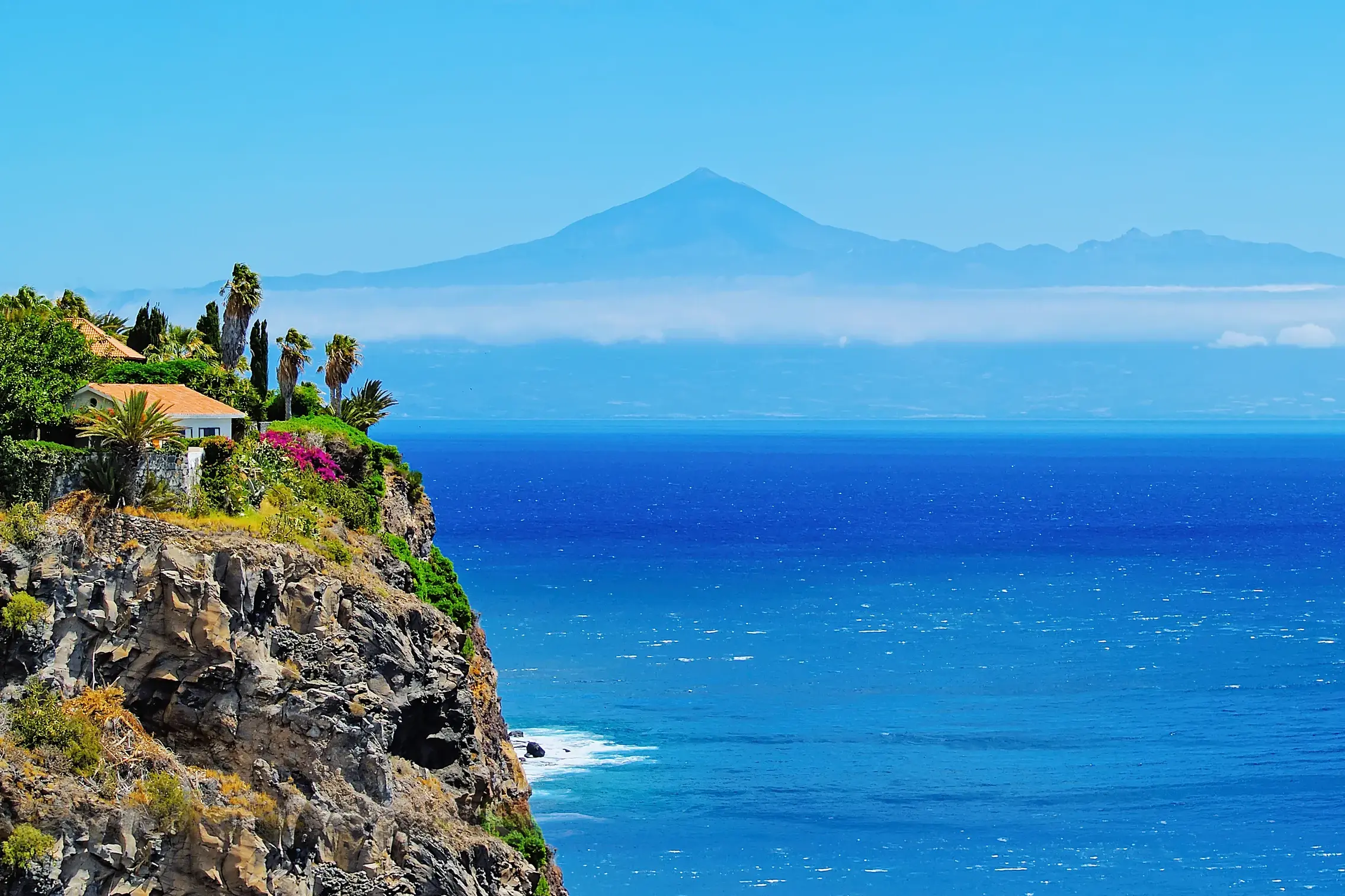 Steile, bewachsene Vulkanklippe an der Küste La Gomeras mit Blick auf den weiten, tiefblauen Atlantik und die Silhouette des Teide auf der Nachbarinsel Teneriffa.