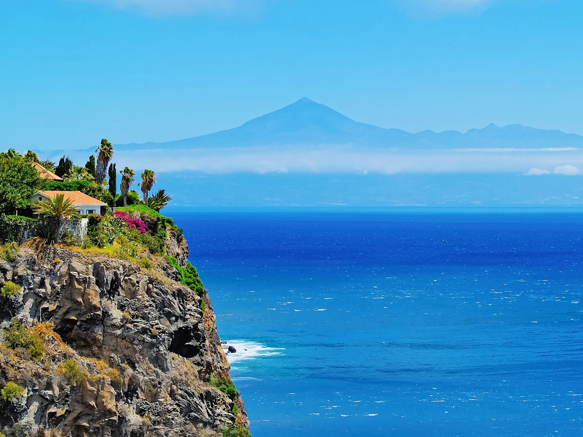 Steile, bewachsene Vulkanklippe an der Küste La Gomeras mit Blick auf den weiten, tiefblauen Atlantik und die Silhouette des Teide auf der Nachbarinsel Teneriffa.