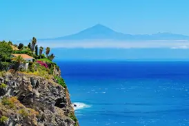 Steile, bewachsene Vulkanklippe an der Küste La Gomeras mit Blick auf den weiten, tiefblauen Atlantik und die Silhouette des Teide auf der Nachbarinsel Teneriffa.
