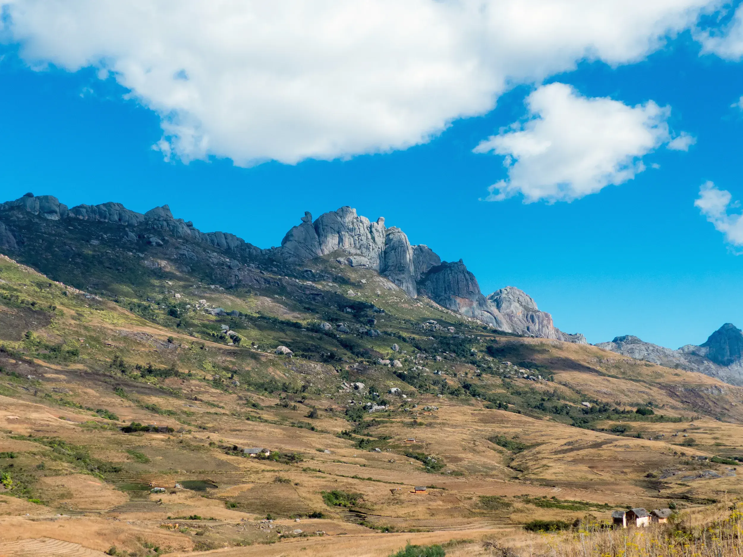 Weite, hügelige Landschaft mit kleinen Hütten vor dem imposanten Andringitra-Massiv in Madagaskar unter blauem Himmel.