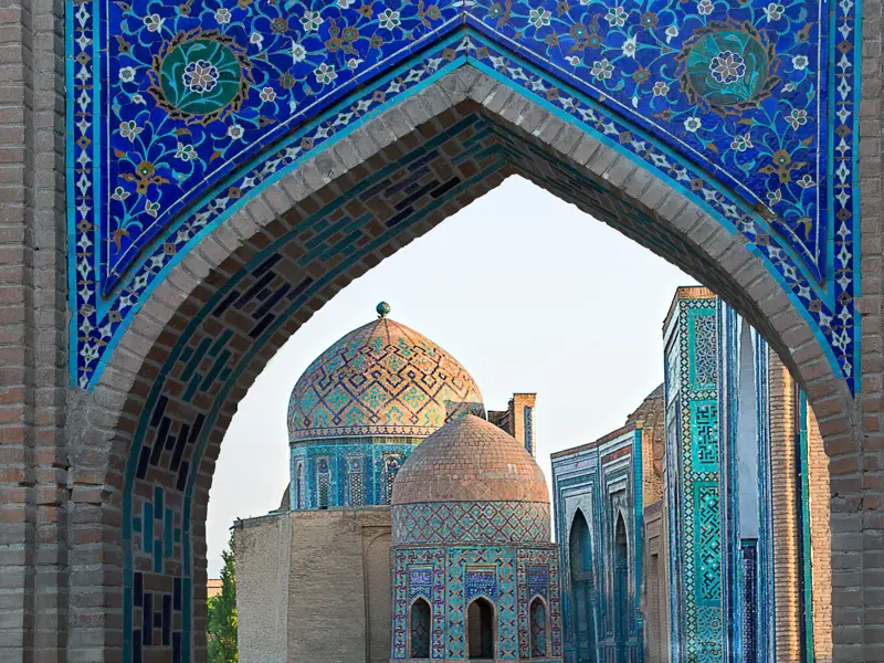 Blick durch ein mit blauen Mosaiken verziertes Tor auf die Kuppeln der Gräberstadt Shohizinda in Samarkand, Usbekistan.