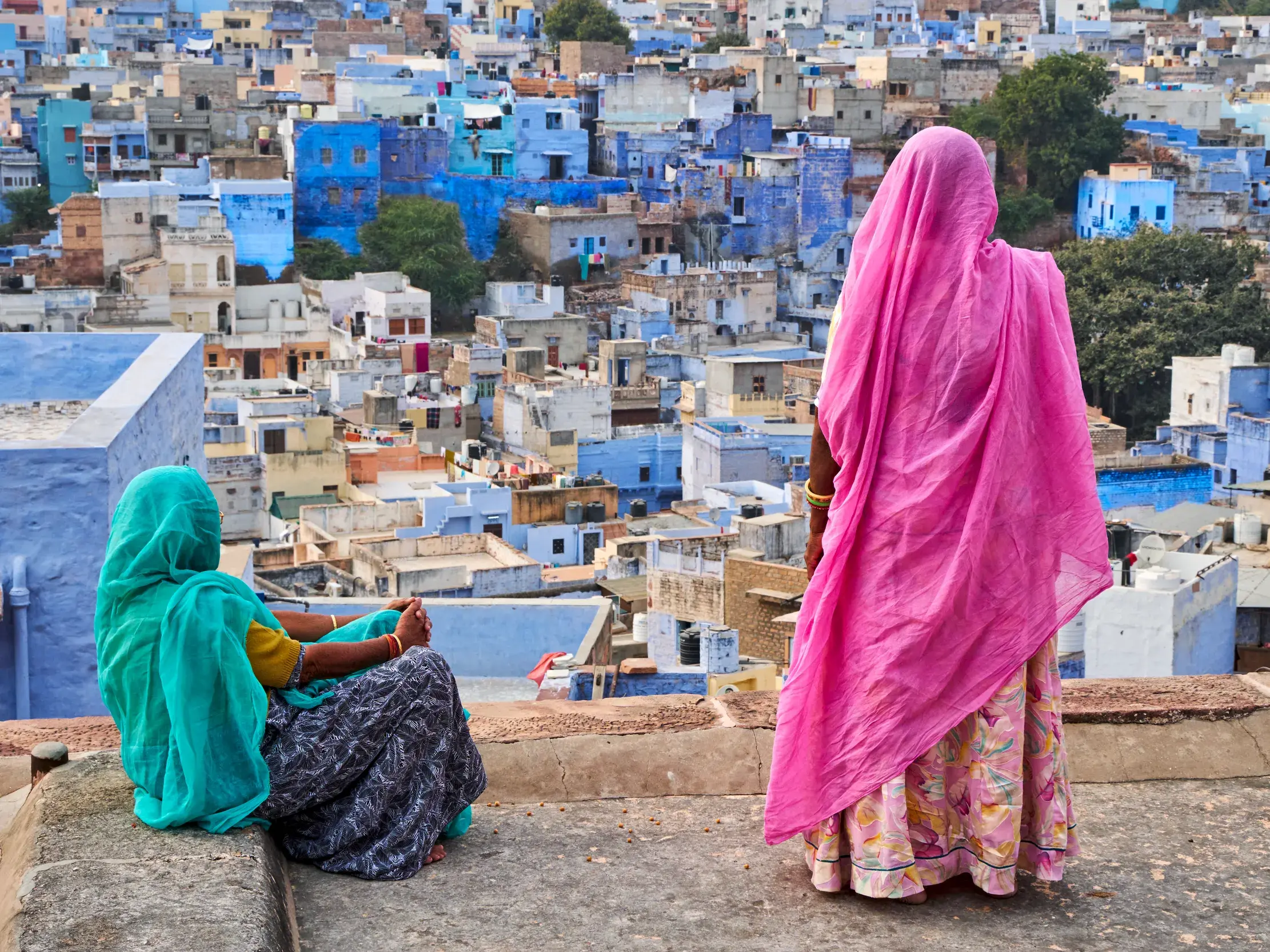 Zwei Frauen in farbenfrohen Saris genießen von einem Dach den Ausblick über die vielen blauen Häuser der Stadt Jodhpur.
