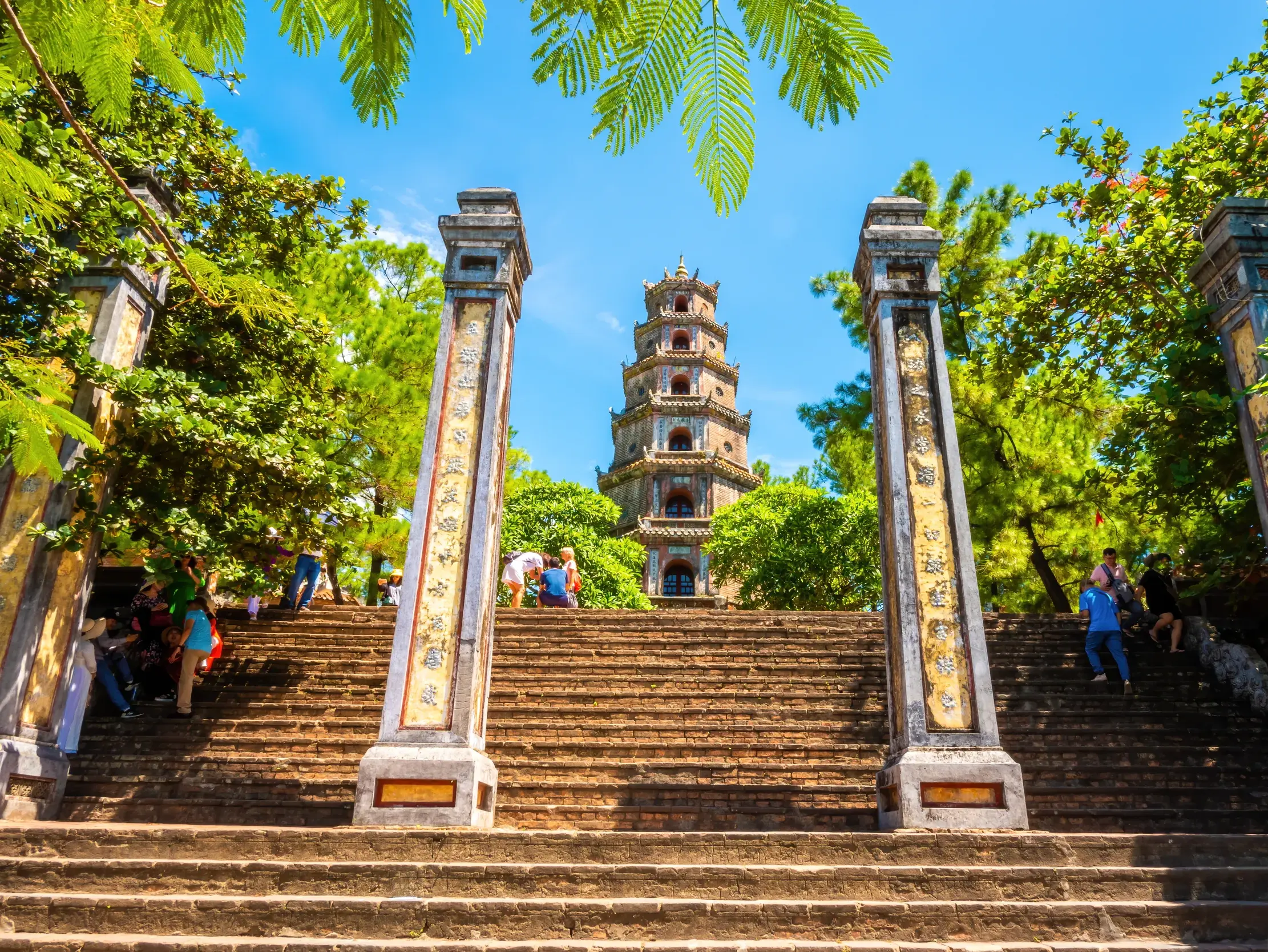 Eine breite Steintreppe führt hinauf zur berühmten Thien-Mu-Pagode in Hue, umrahmt von verzierten Säulen und grünen Bäumen.