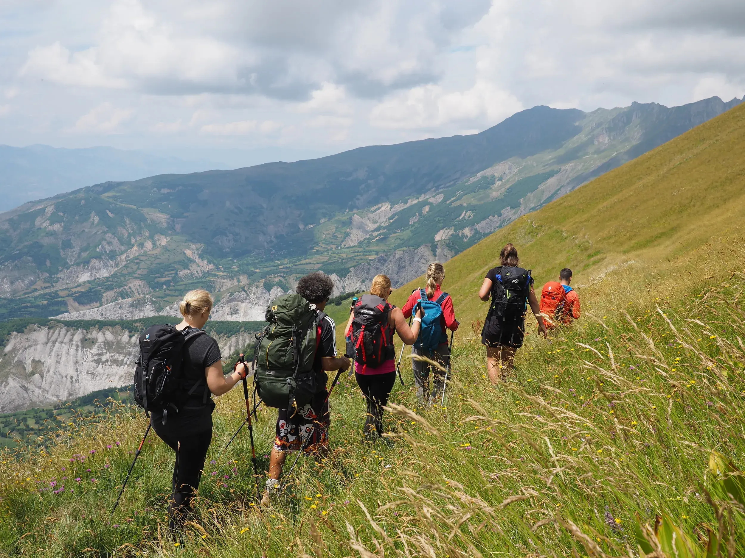 Eine Reisegruppe mit Trekkingrucksäcken wandert auf einem Pfad durch eine hochalpine Wiese im albanischen Korab-Gebirge.