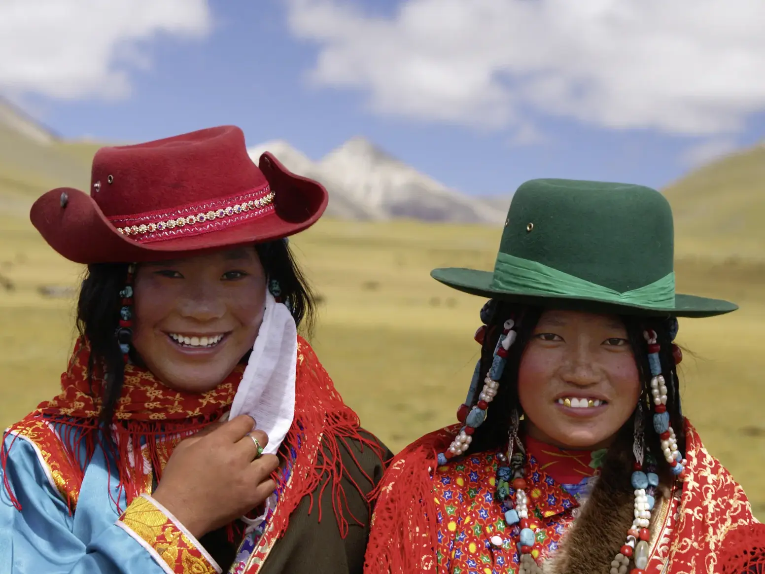Zwei junge tibetische Frauen in traditioneller Kleidung und Hüten lächeln herzlich vor der Bergkulisse der Hochebene Tibets.