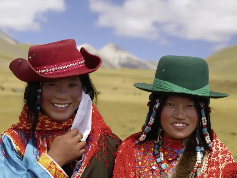 Zwei junge tibetische Frauen in traditioneller Kleidung und Hüten lächeln herzlich vor der Bergkulisse der Hochebene Tibets.