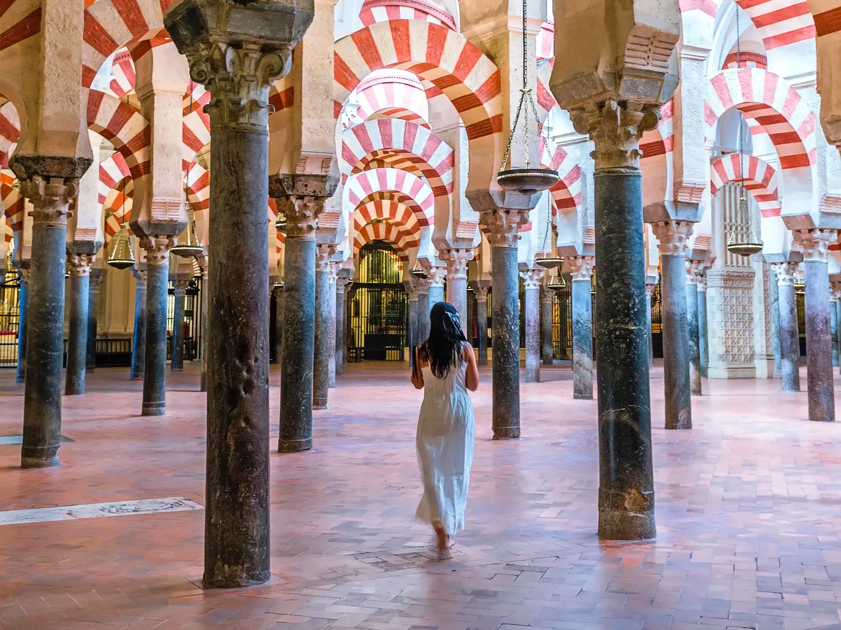 Frau in weißem Kleid läuft durch die endlosen Säulenreihen mit rot-weißen Hufeisenbögen in der Mezquita in Córdoba.