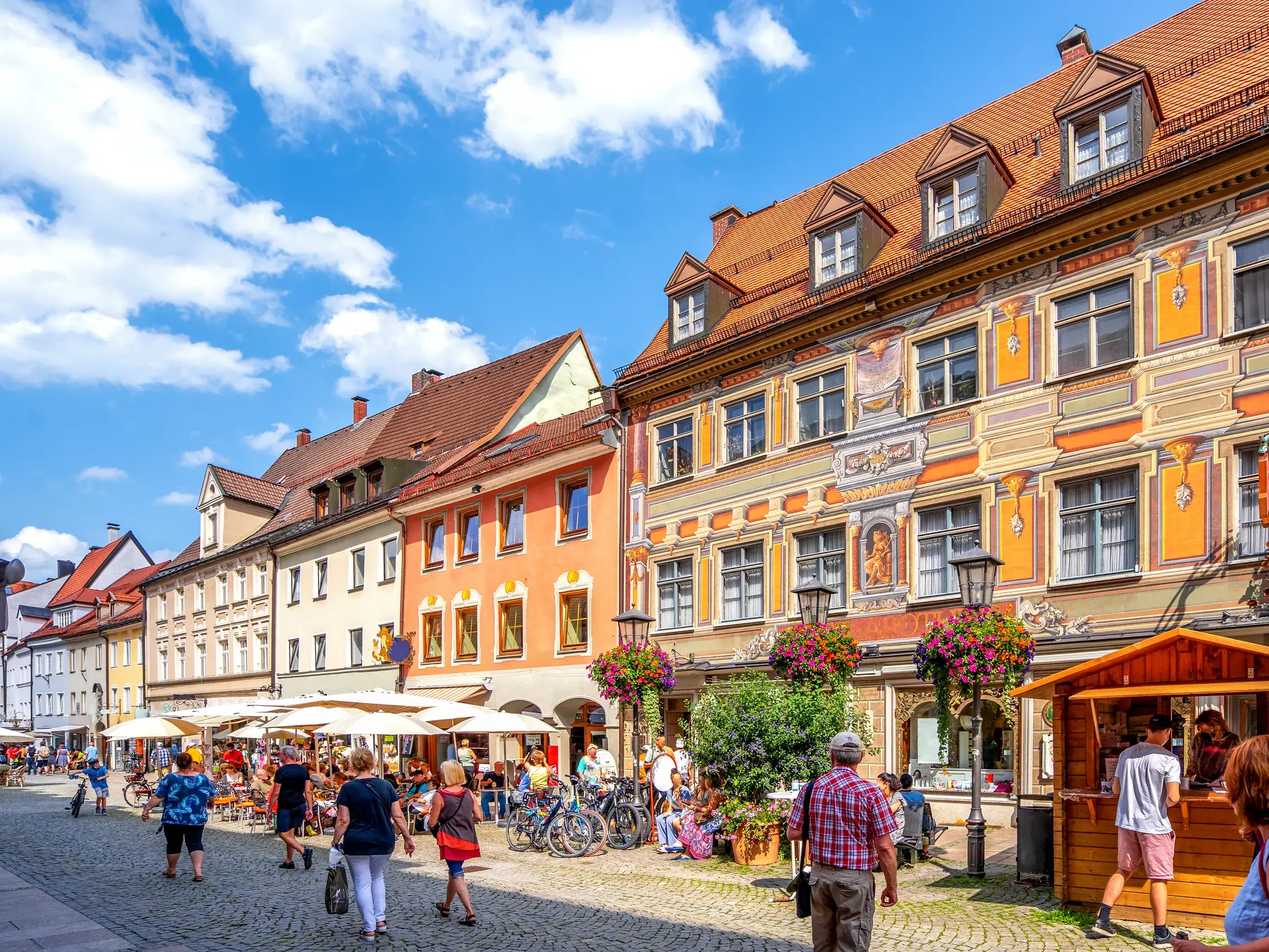 Sonnige Gasse in der Altstadt von Füssen mit bunten Häuserfassaden, belebten Straßencafés und flanierenden Menschen.