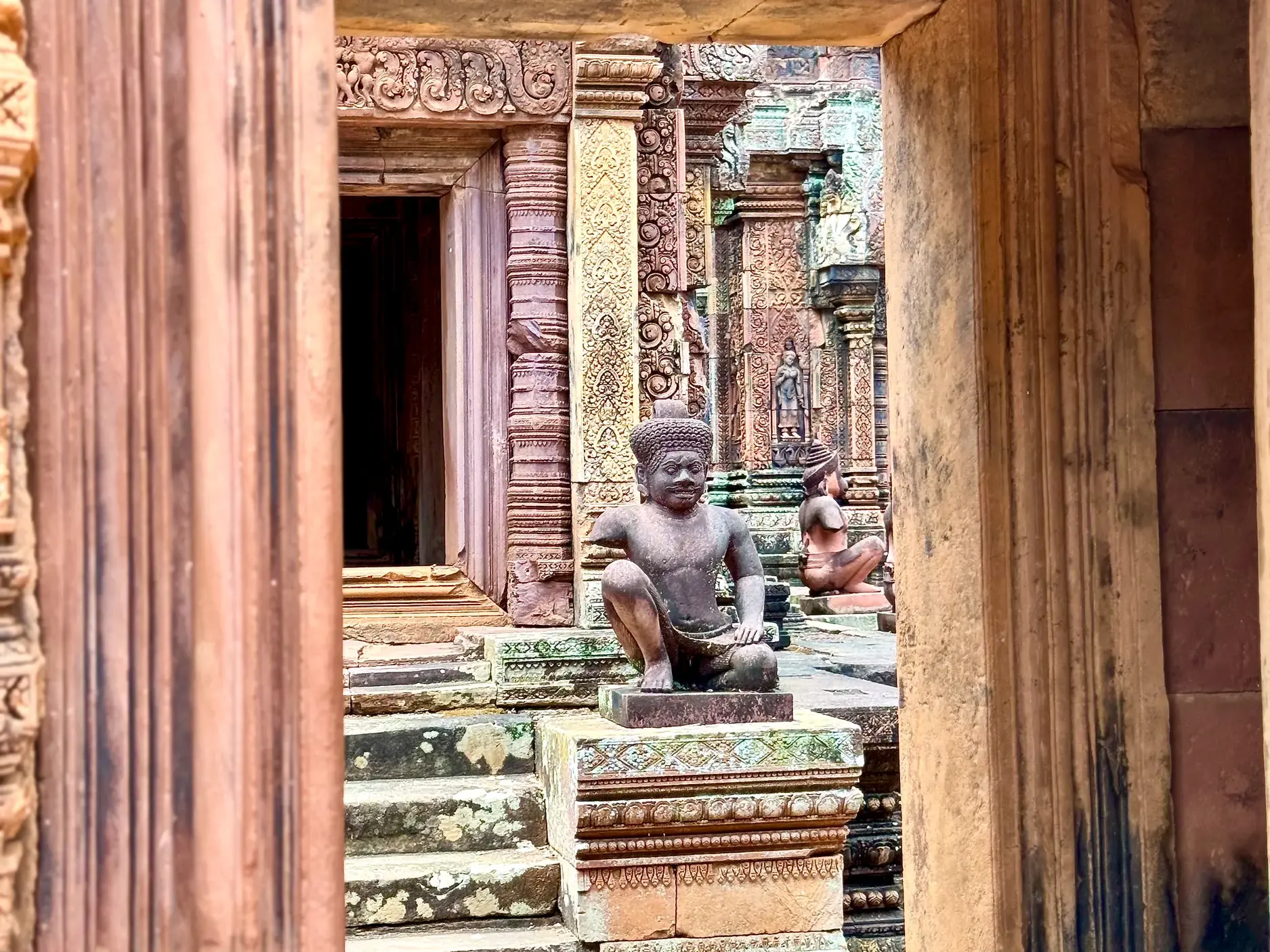 Hockende Wächterstatue aus rotem Sandstein vor kunstvollen Reliefs im Banteay Srei Tempel in Angkor, Kambodscha.