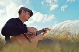 Ein Musiker mit Schiebermütze spielt auf seiner Gitarre melancholische Lieder in der weiten, grasbewachsenen Landschaft Irlands.