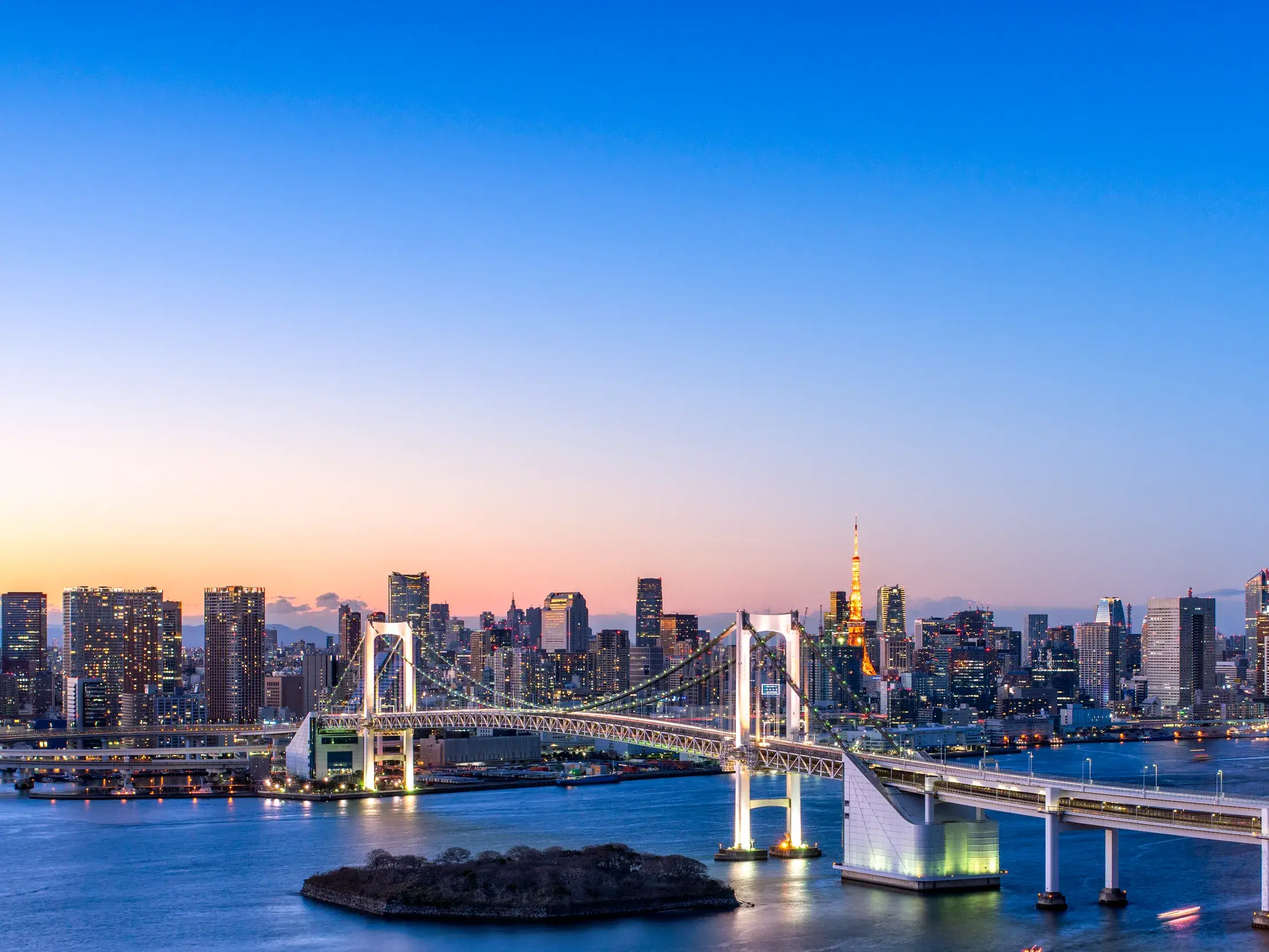 Panoramablick von Odaiba auf die beleuchtete Rainbow Bridge und die Skyline von Tokio in der Abenddämmerung.