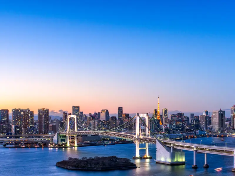 Panoramablick von Odaiba auf die beleuchtete Rainbow Bridge und die Skyline von Tokio in der Abenddämmerung.