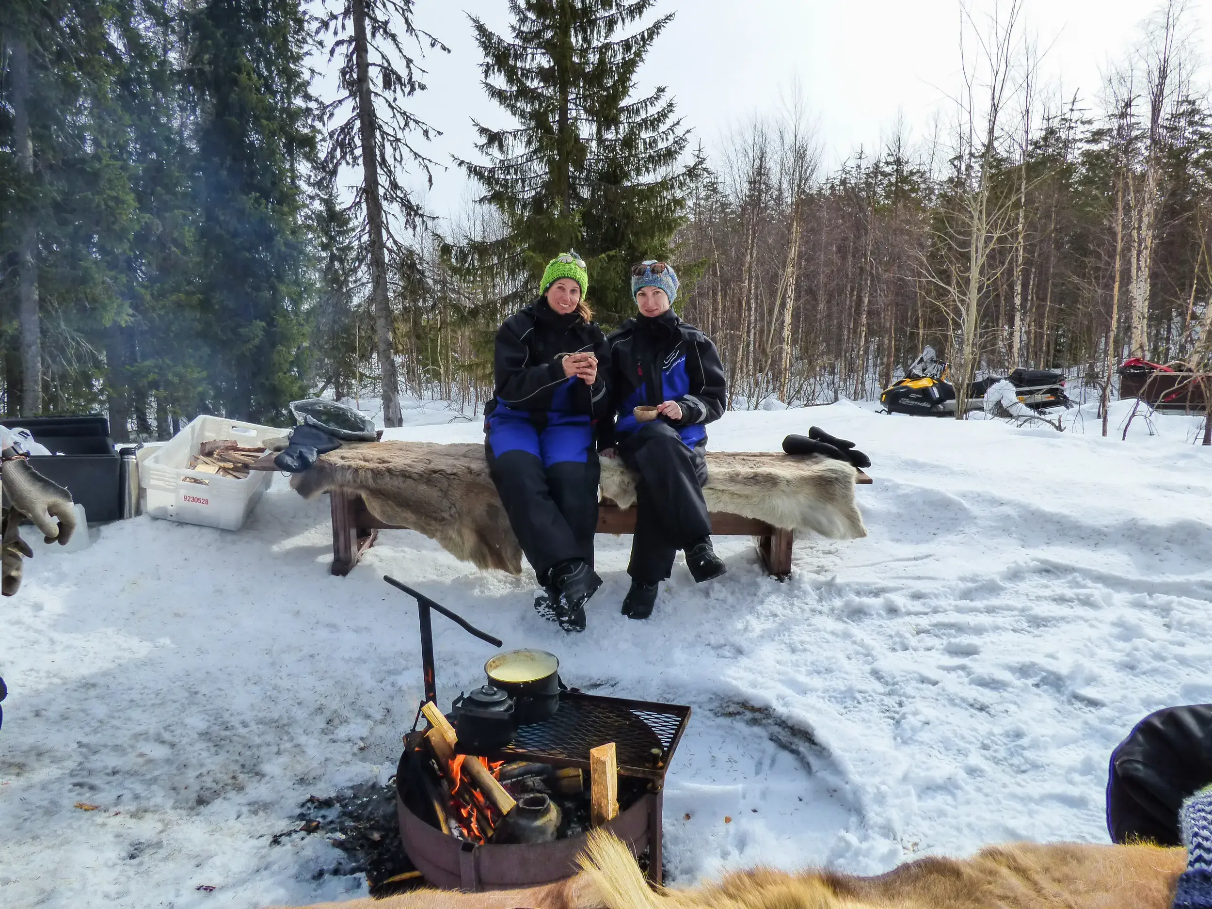 Zwei Frauen einer Gruppenreise entspannen mit heißen Getränken am Lagerfeuer während einer Pause in der finnischen Schneelandschaft.