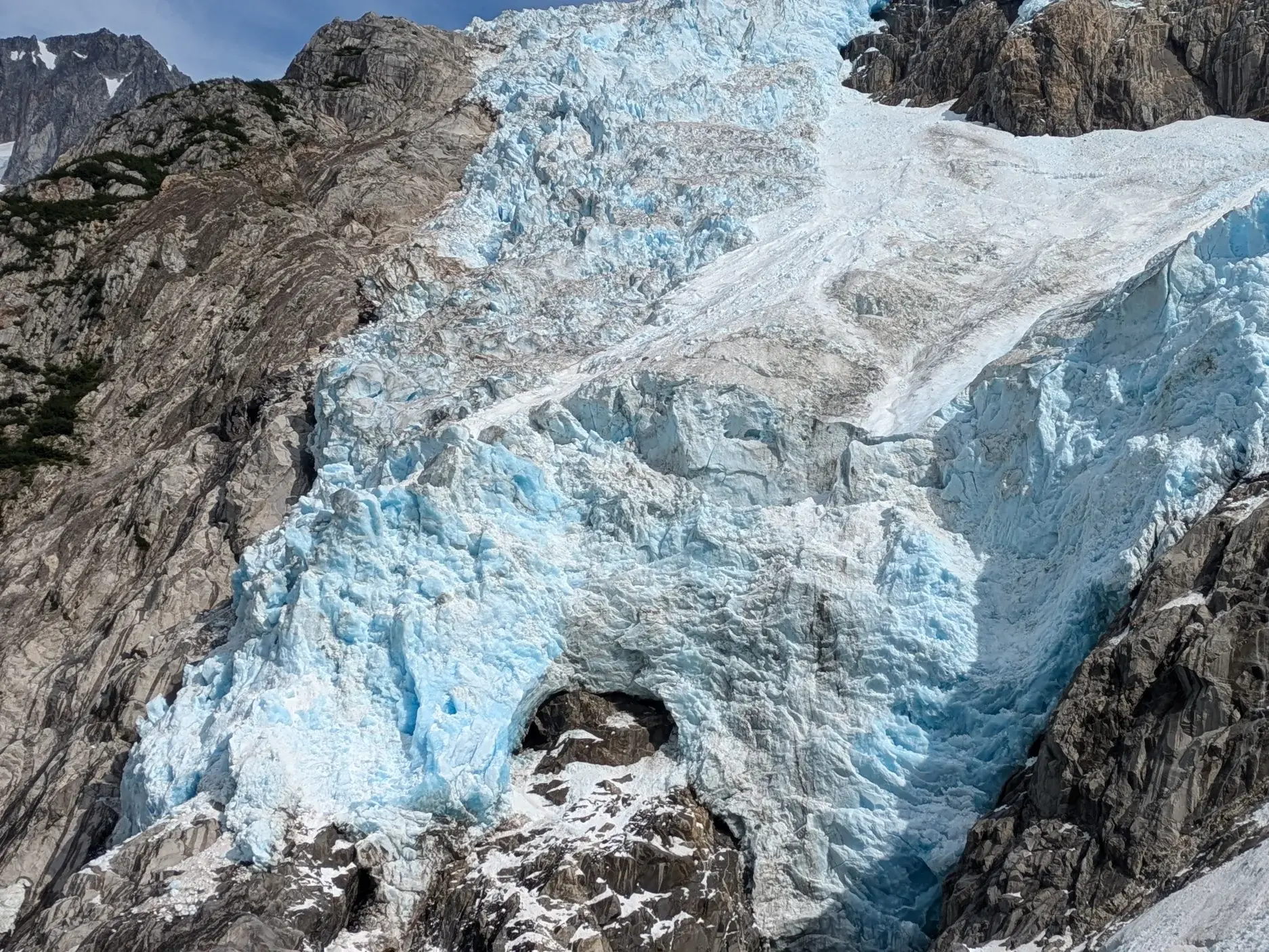 Nahaufnahme einer bläulich schimmernden Gletscherzunge mit Eishöhle an einer Felswand im Kenai Fjords Nationalpark in Alaska.