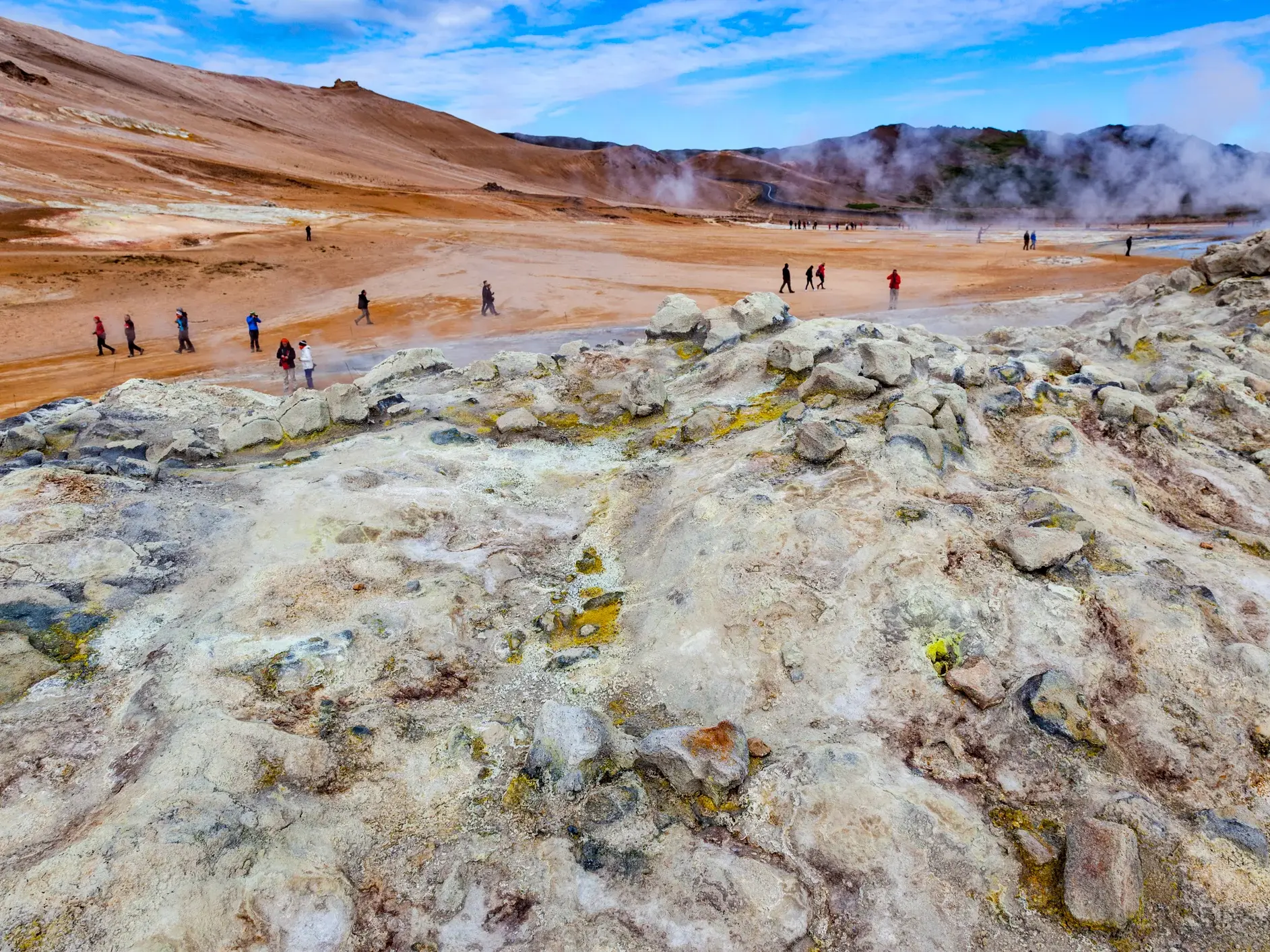 Reisende wandern durch das weite, dampfende Geothermalgebiet am Myvatn mit seinen farbenfrohen Schwefelablagerungen.