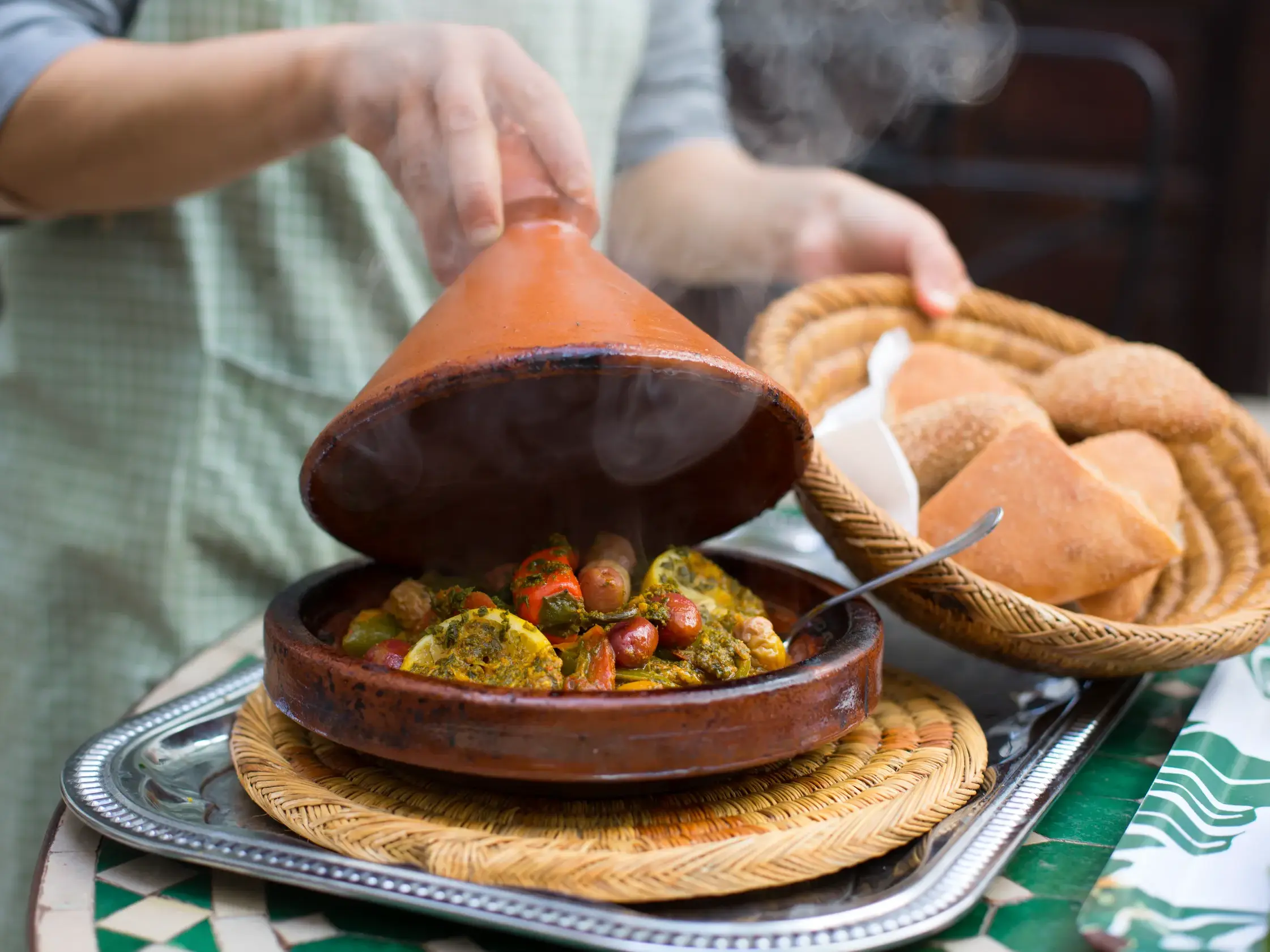 Eine Person lüftet den Deckel einer dampfenden Gemüse-Tajine während eines Kochkurses in Marrakesch.