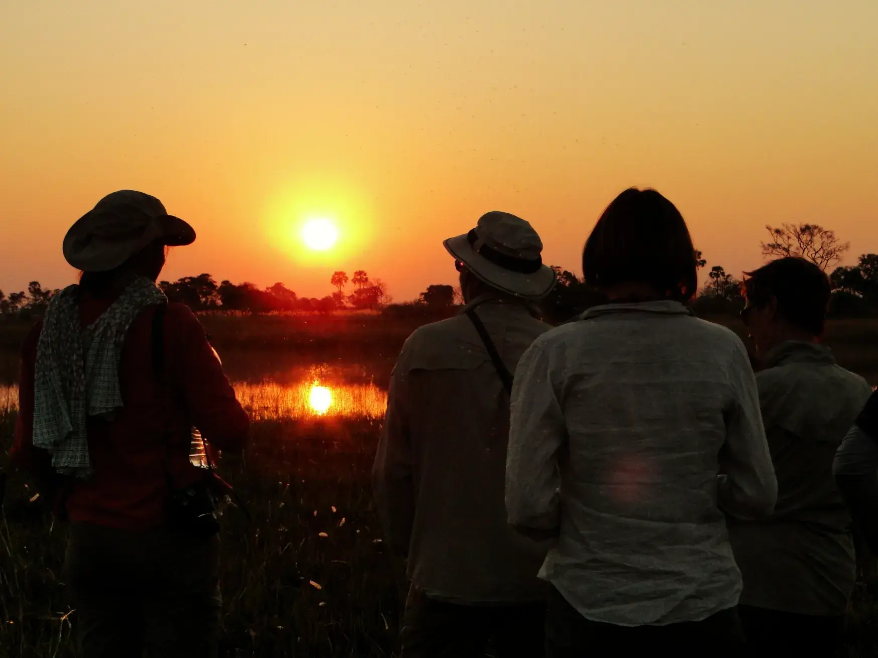 Eine kleine Reisegruppe in Outdoor-Kleidung beobachtet den goldenen Sonnenuntergang über einer Wasserstelle in Südafrika.