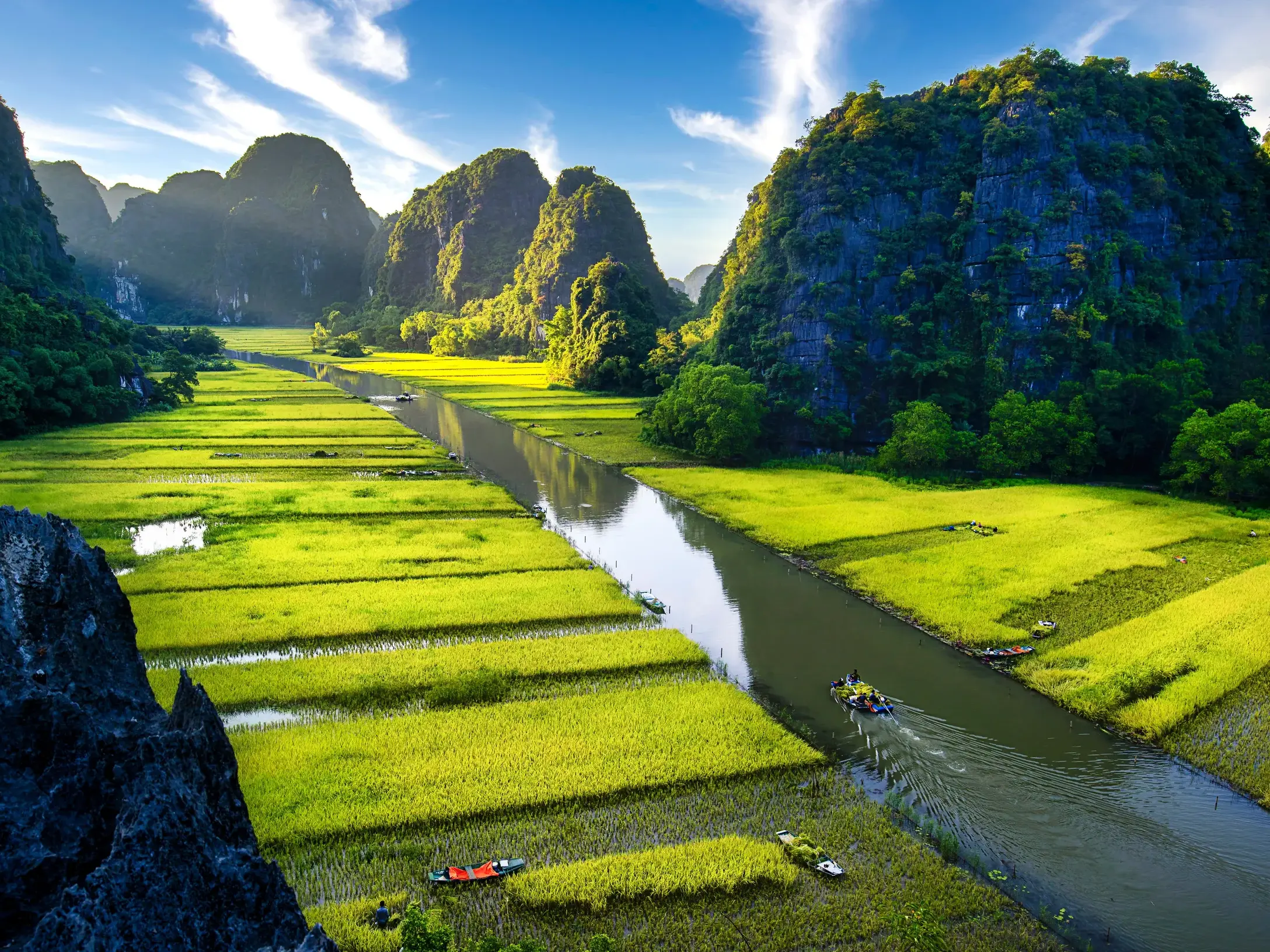 Panoramablick auf den Ngo Dong Fluss in Tam Coc mit grünen Reisfeldern, Karstbergen und kleinen Booten bei Sonnenschein.