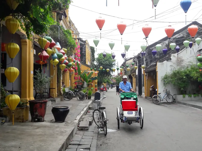 Ein Mann fährt mit einer traditionellen Cyclo-Rikscha durch die mit bunten Lampions dekorierte Altstadt von Hoi An.