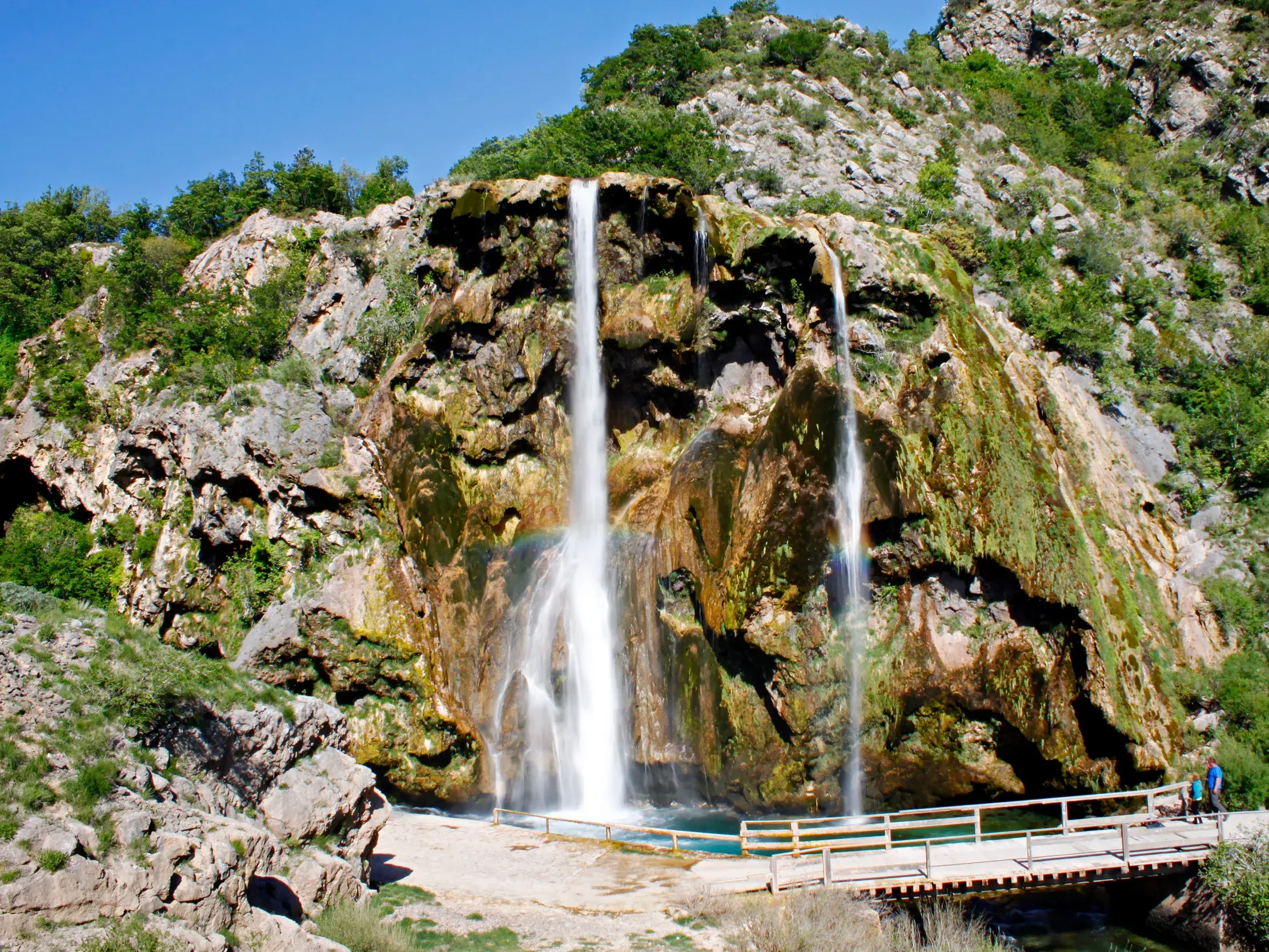 Wanderer auf einer Holzbrücke vor dem imposanten Wasserfall des Krupa-Flusses, der in Kroatien über moosige Felsen stürzt.