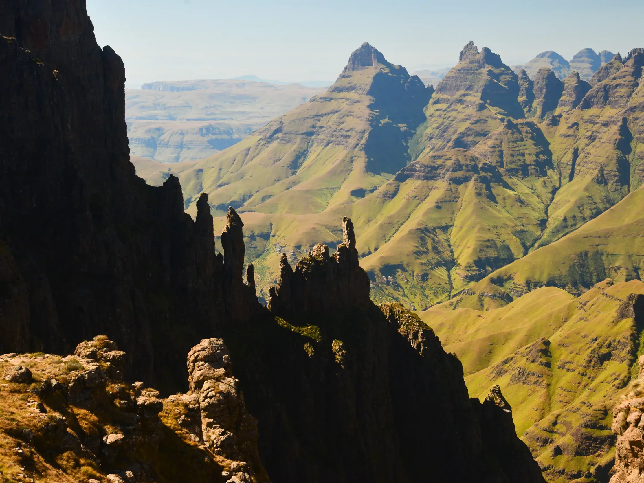 Raue Felsformationen im Schatten mit Blick auf die sonnenbeschienene, grüne Berglandschaft der Drakensberge in Südafrika.