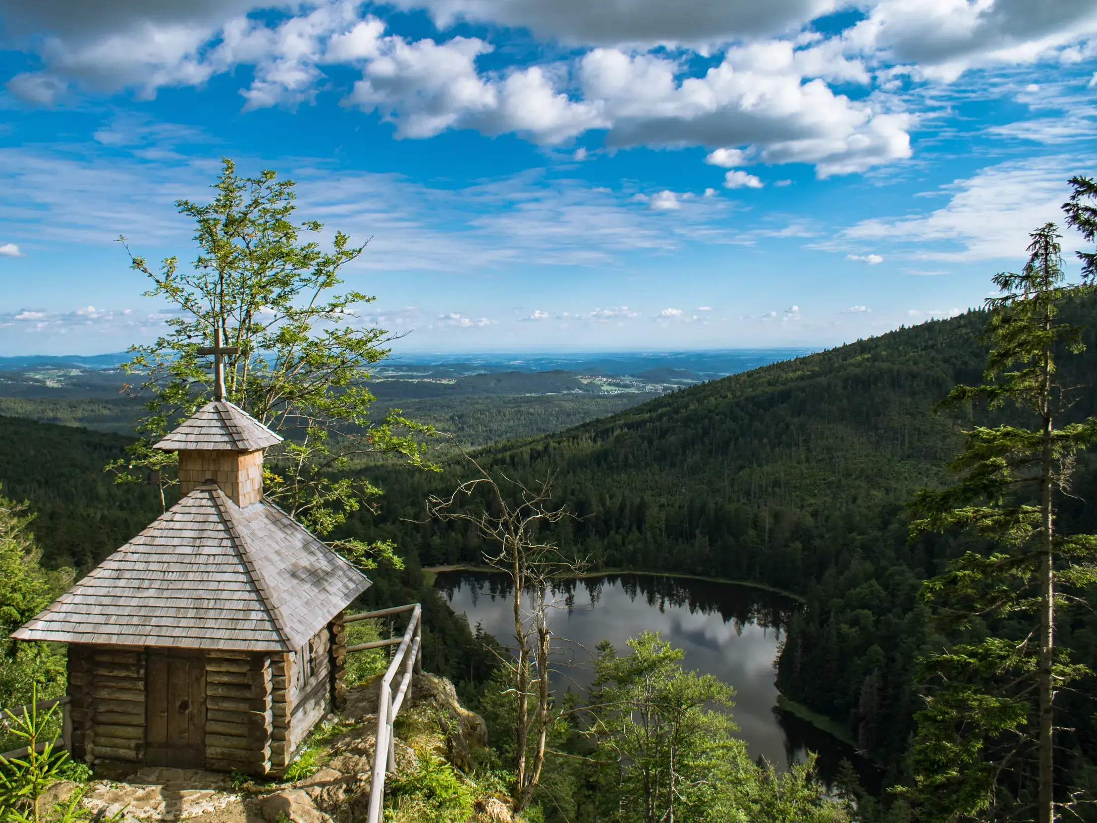Kleine Holzkapelle auf einem Felsvorsprung mit Panoramablick über einen Bergsee und eine weite, grüne Waldlandschaft.