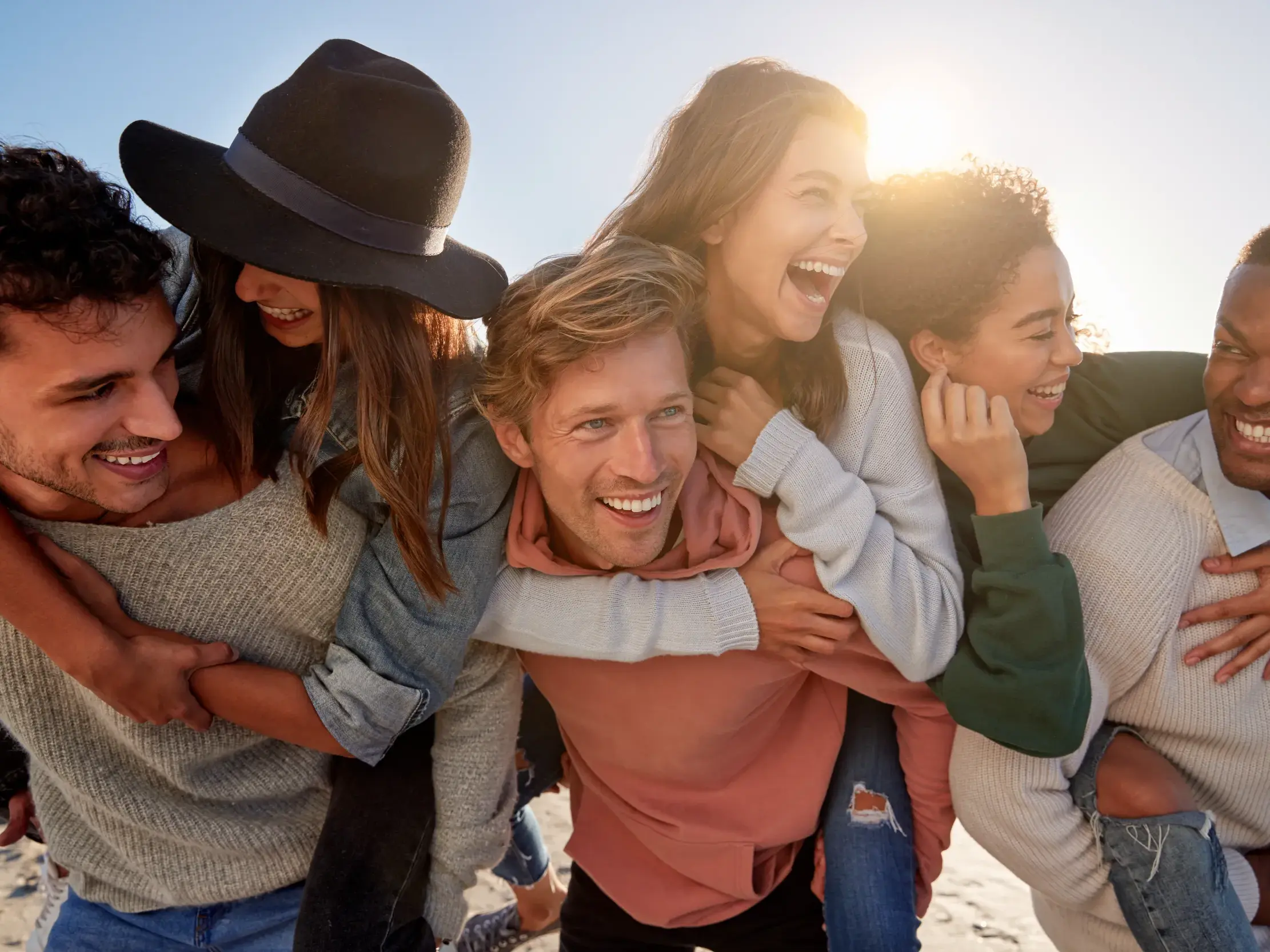 Junge Leute einer Reisegruppe lachen ausgelassen, während sie sich bei strahlendem Sonnenschein am Strand Huckepack tragen.