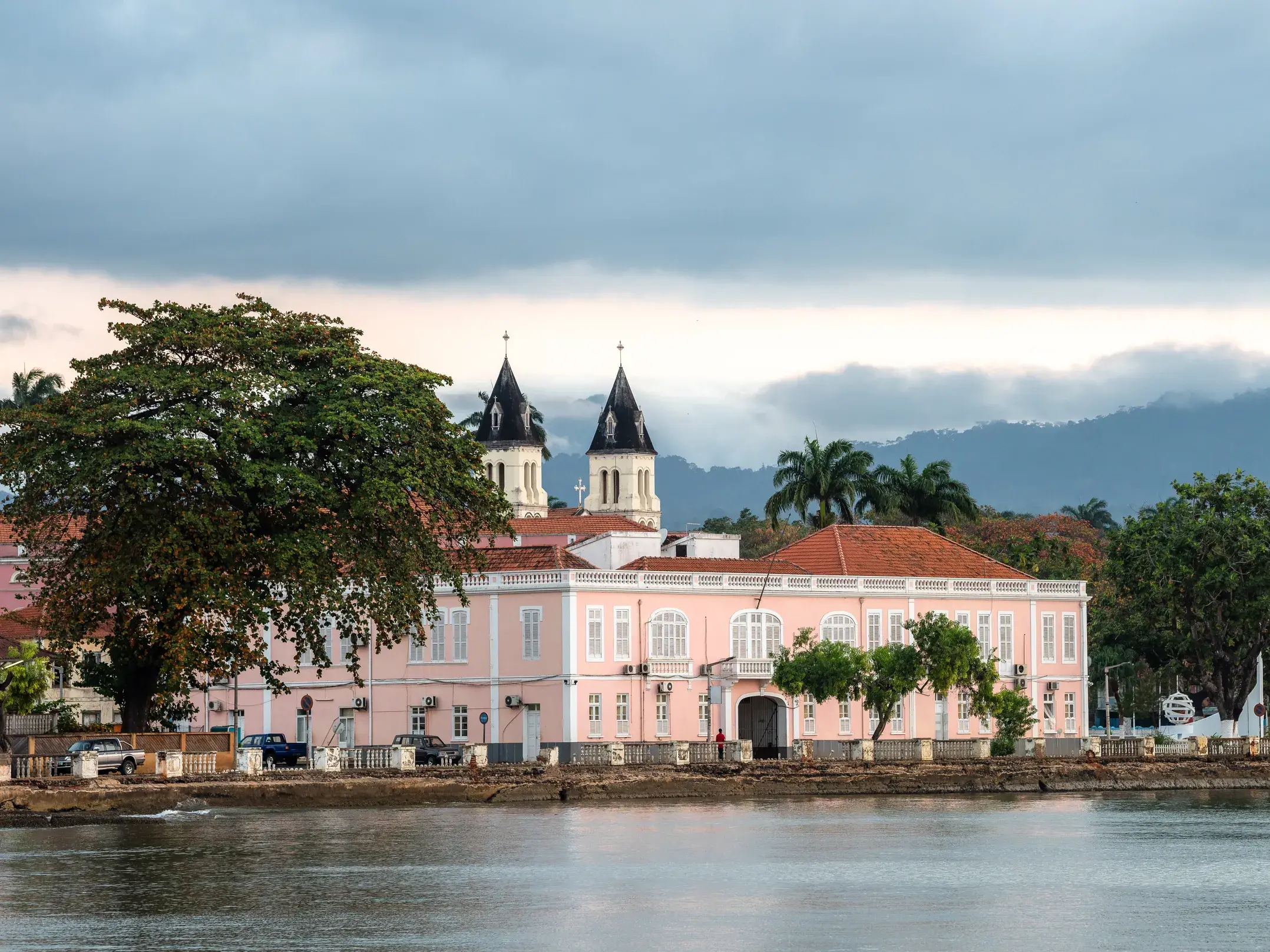 Rosafarbenes Kolonialgebäude an der Uferpromenade von São Tomé, dahinter die Türme der Kathedrale und grüne Berge.