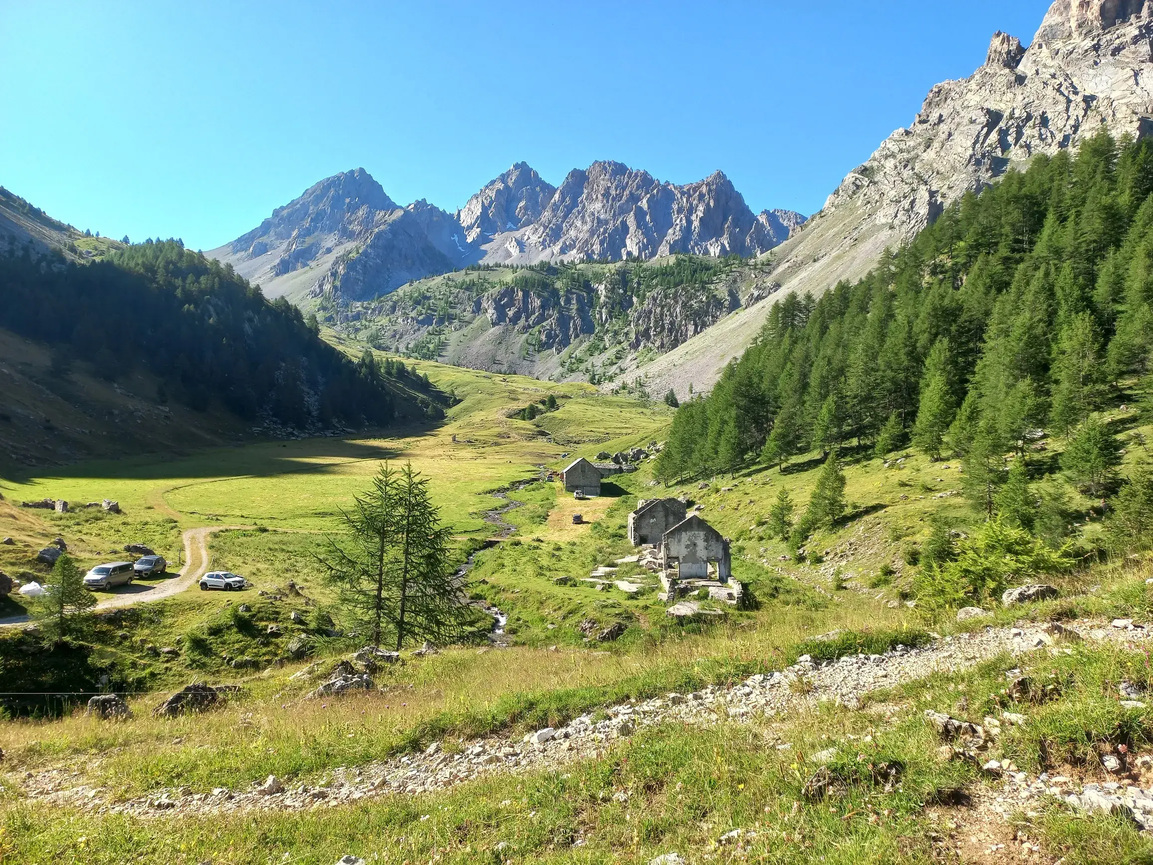 Sonniges Bergpanorama im Maira-Tal im Piemont mit grünen Wiesen, alten Steinhütten und schroffen Alpengipfeln im Hintergrund.