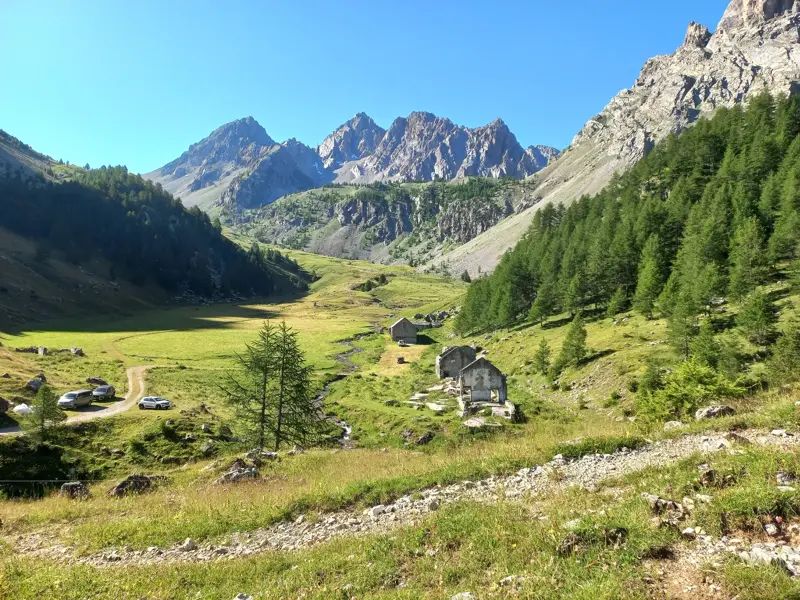Sonniges Bergpanorama im Maira-Tal im Piemont mit grünen Wiesen, alten Steinhütten und schroffen Alpengipfeln im Hintergrund.
