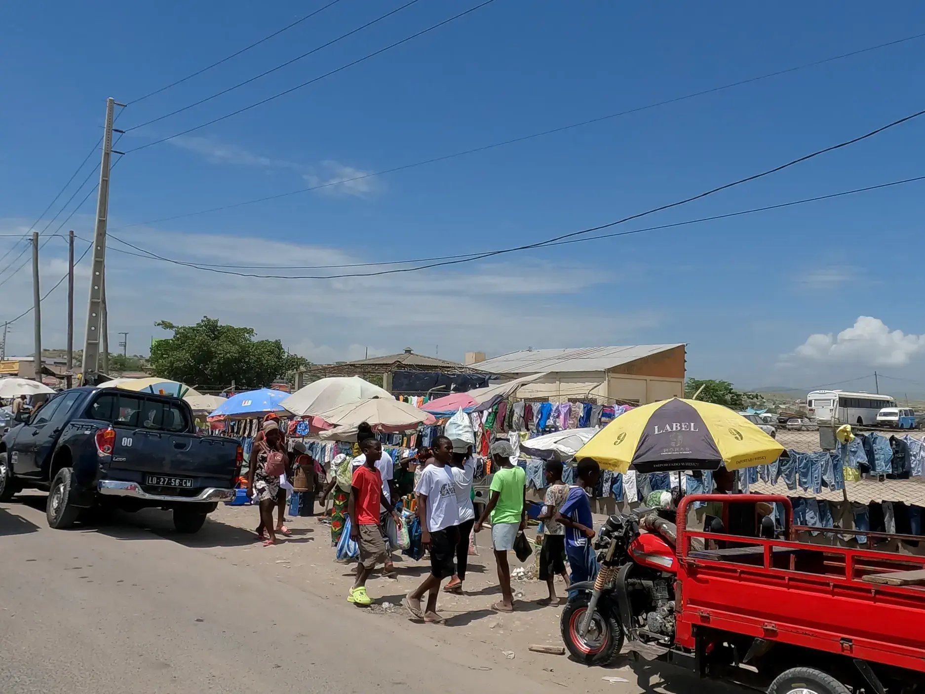 Authentische Straßenszene auf einem belebten Markt in Angola mit Einheimischen, bunten Verkaufsständen und einem Pickup am Straßenrand.
