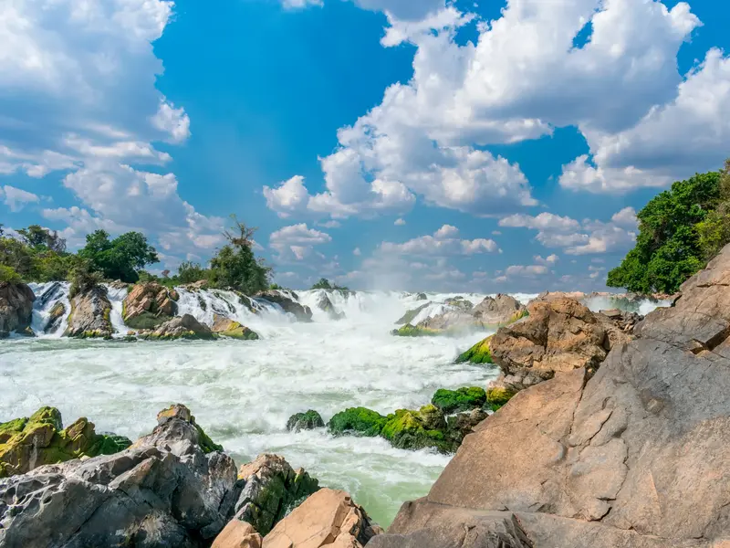 Tosende Wassermassen des Khong Phapheng Wasserfalls in Südlaos, umgeben von moosbewachsenen Felsen unter blauem Wolkenhimmel.