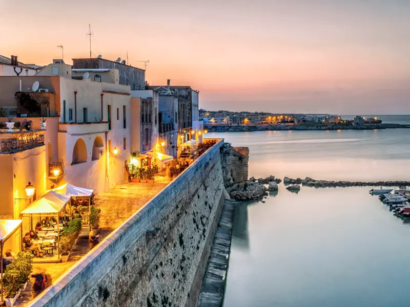 Stimmungsvolle Abenddämmerung in Otranto mit Blick auf die beleuchtete Altstadt, die Bastionen und den ruhigen Hafen an der Adria.