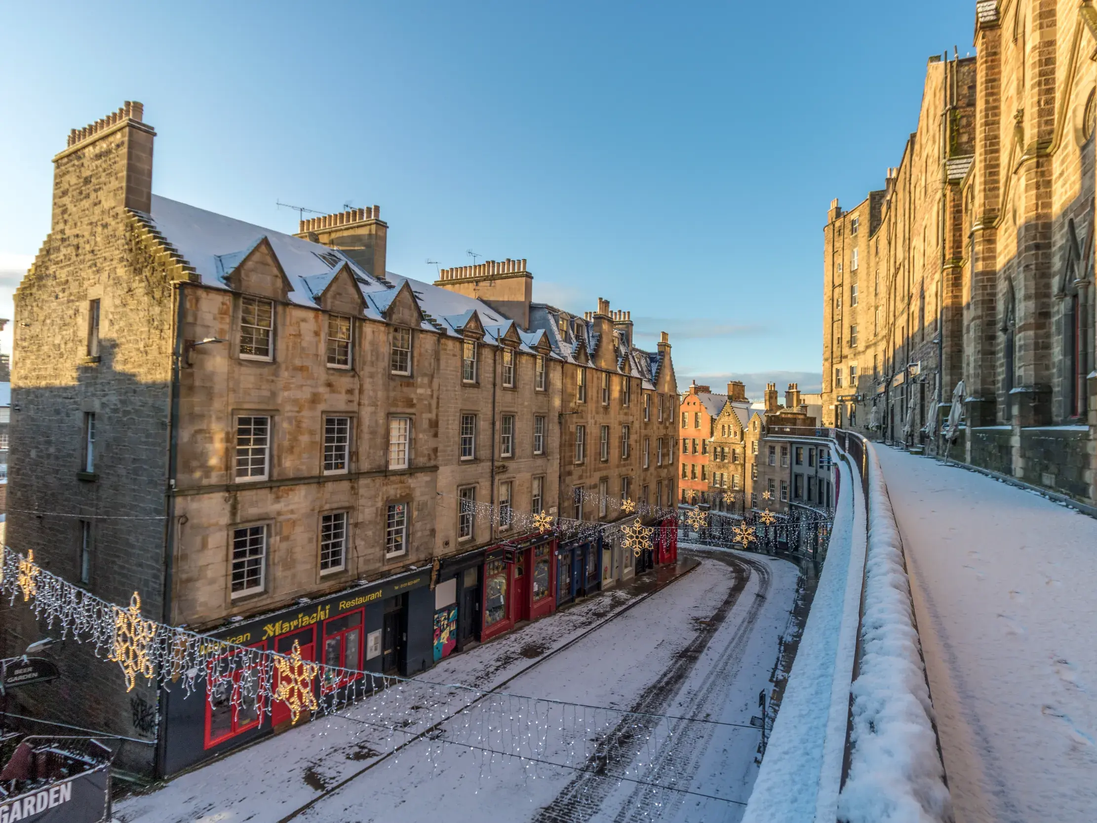 Blick auf die schneebedeckte, historische Altstadt von Edinburgh mit festlicher Beleuchtung an einem sonnigen Wintertag.