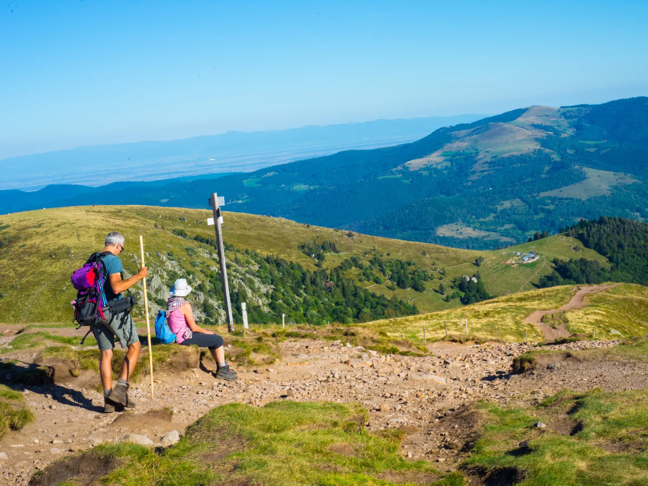 Zwei Wanderer mit Rucksäcken bei einer Rast auf dem steinigen Vogesenkamm mit Panoramablick über die Hügel am Hohneck.