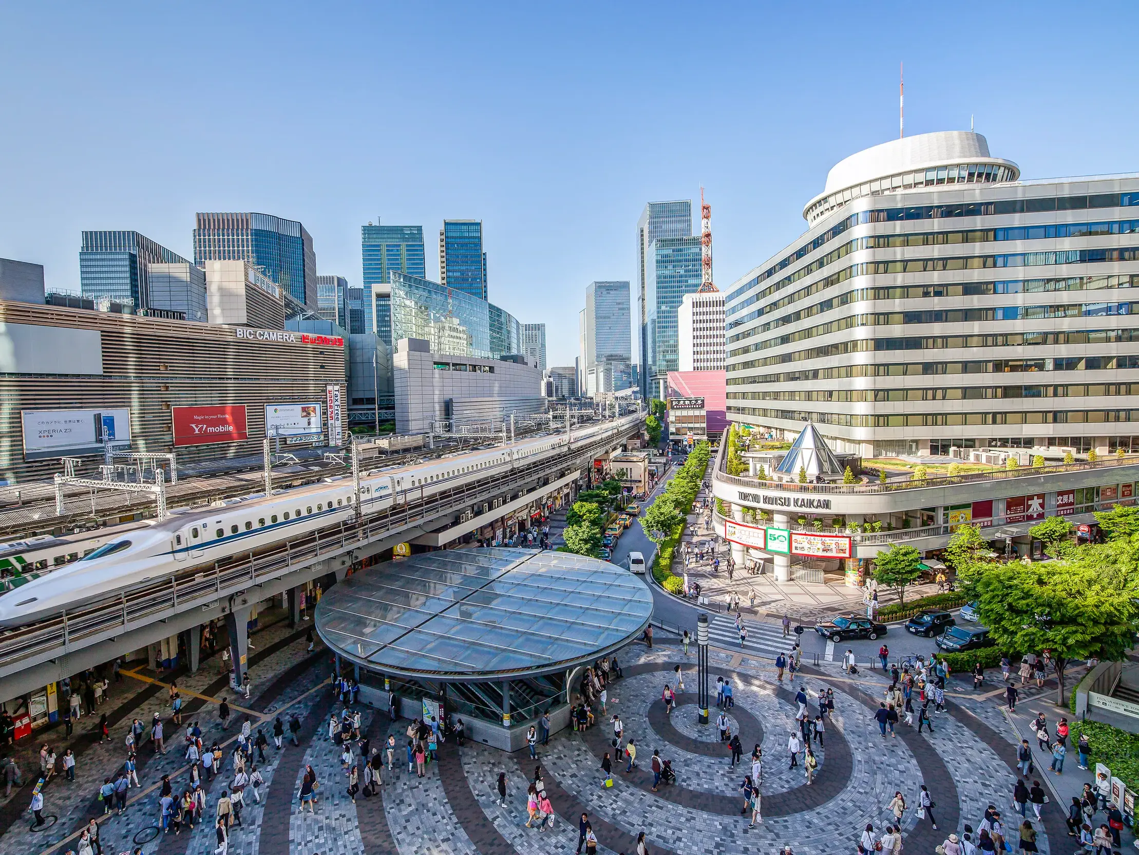 Ein Shinkansen-Schnellzug fährt durch die beeindruckende Hochhausschlucht von Tokio, über einen belebten Platz nahe Yurakucho.