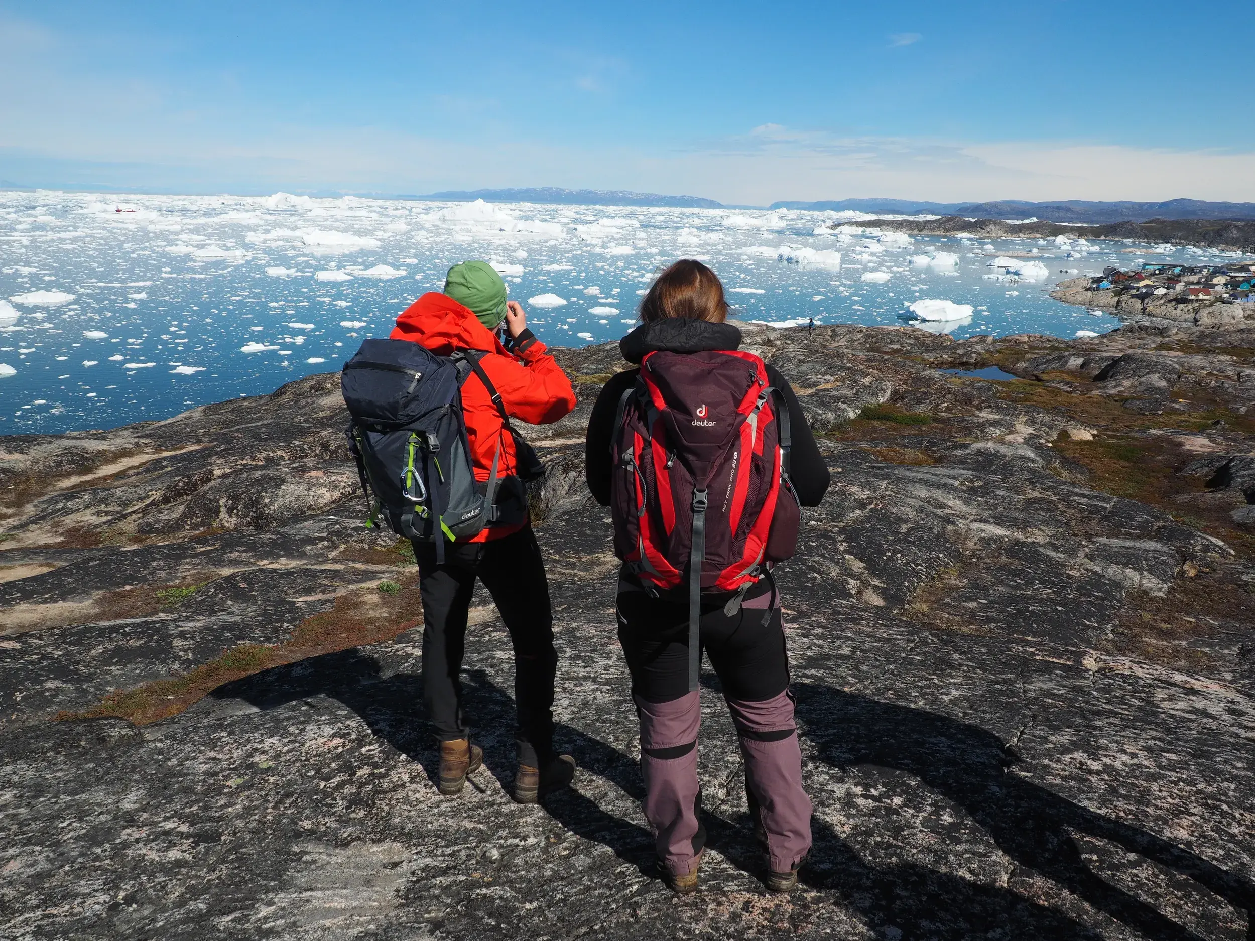 Zwei Wanderer mit Trekking-Rucksäcken blicken von einem Felsen auf die Diskobucht voller Eisberge bei Ilulissat in Grönland.