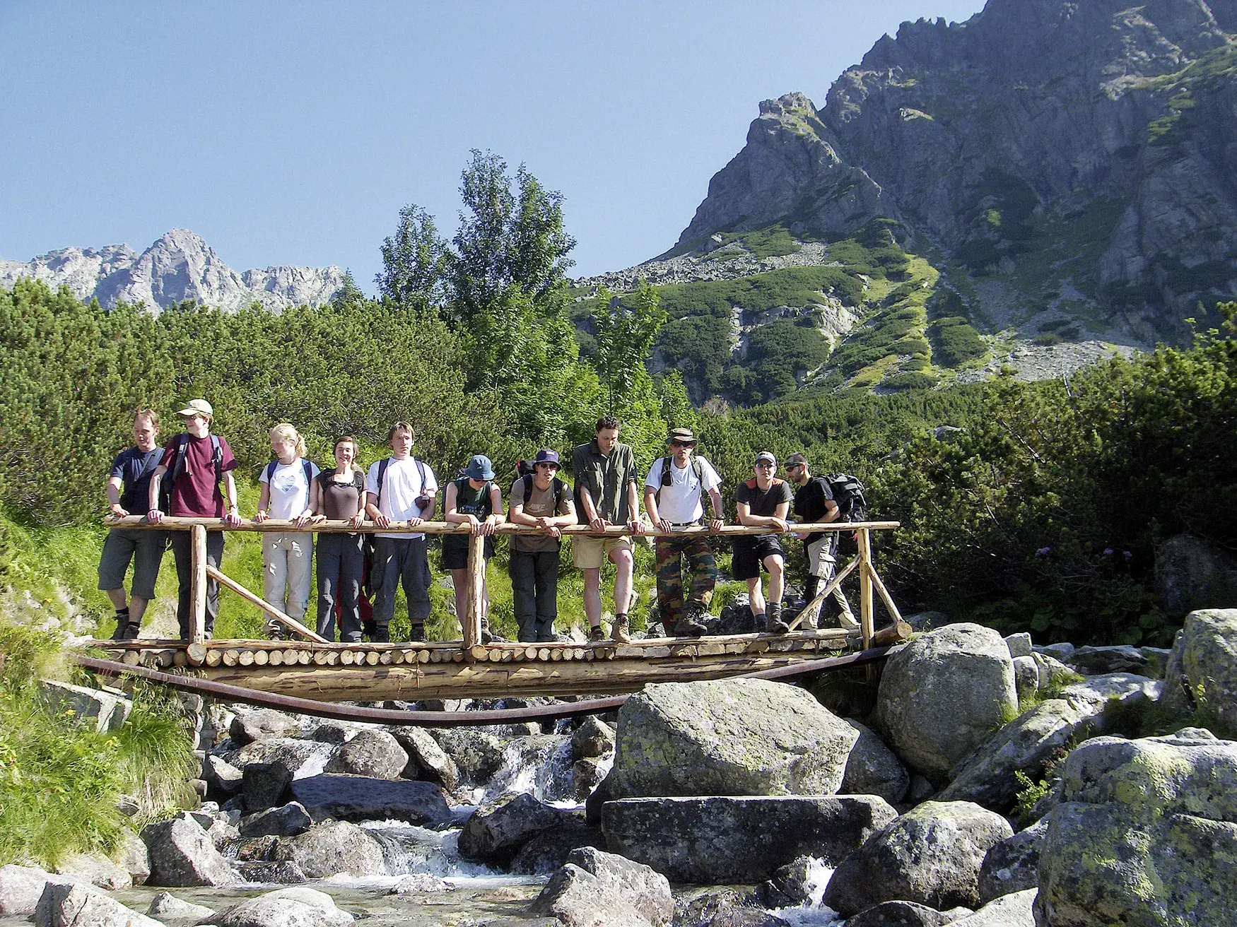 Eine Hauser-Wandergruppe pausiert für ein Foto auf einer Holzbrücke über einem Gebirgsbach in der Berglandschaft der Hohen Tatra.
